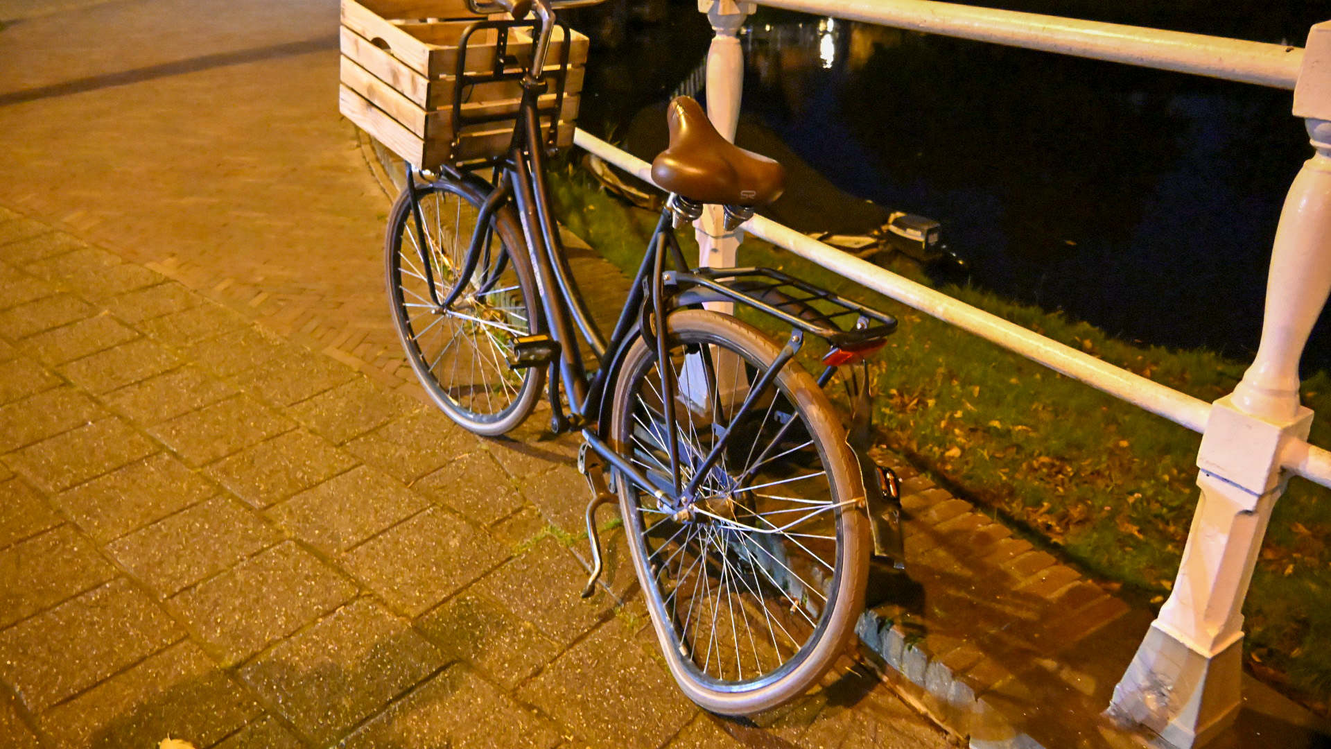 Donkergekleurde fiets met een houten krat op de bagagedrager, geparkeerd langs een brugleuning bij nacht.