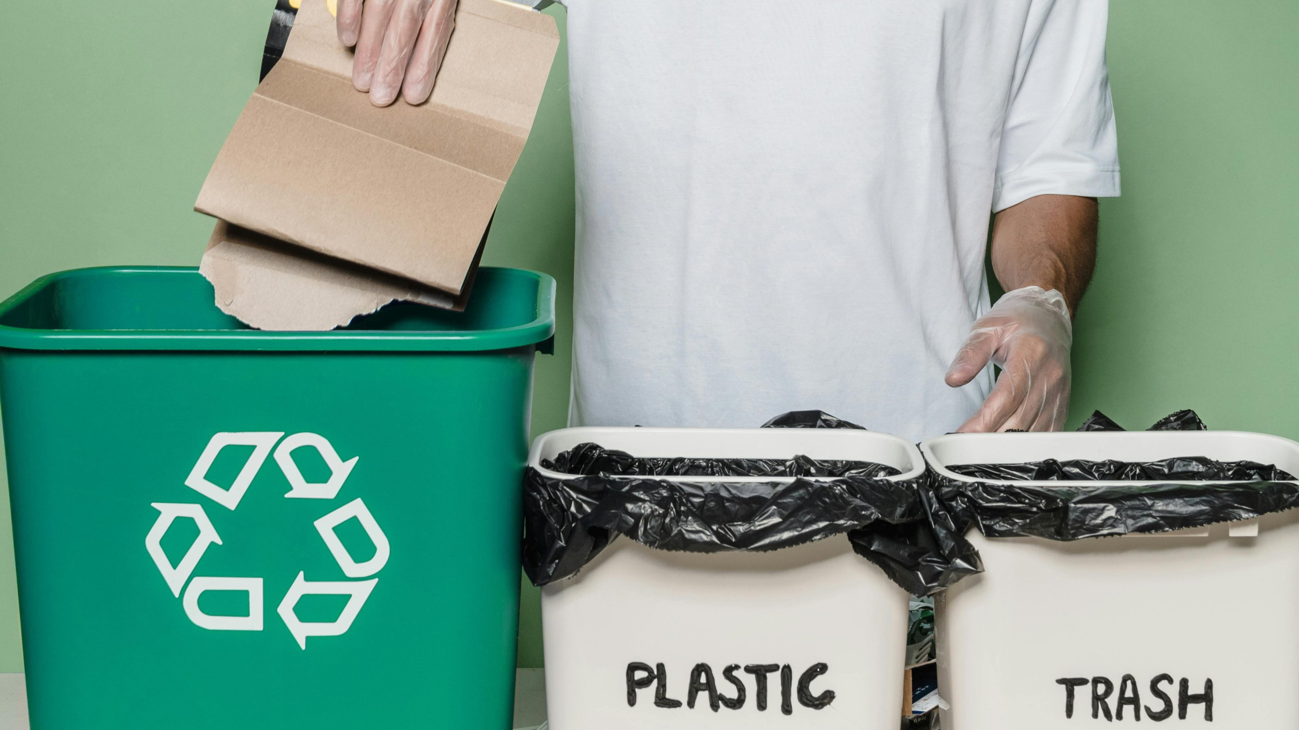 Een persoon gooit karton in een groene prullenbak met het recyclinglogo, naast twee witte bakken gemarkeerd met 'PLASTIC' en 'TRASH'.