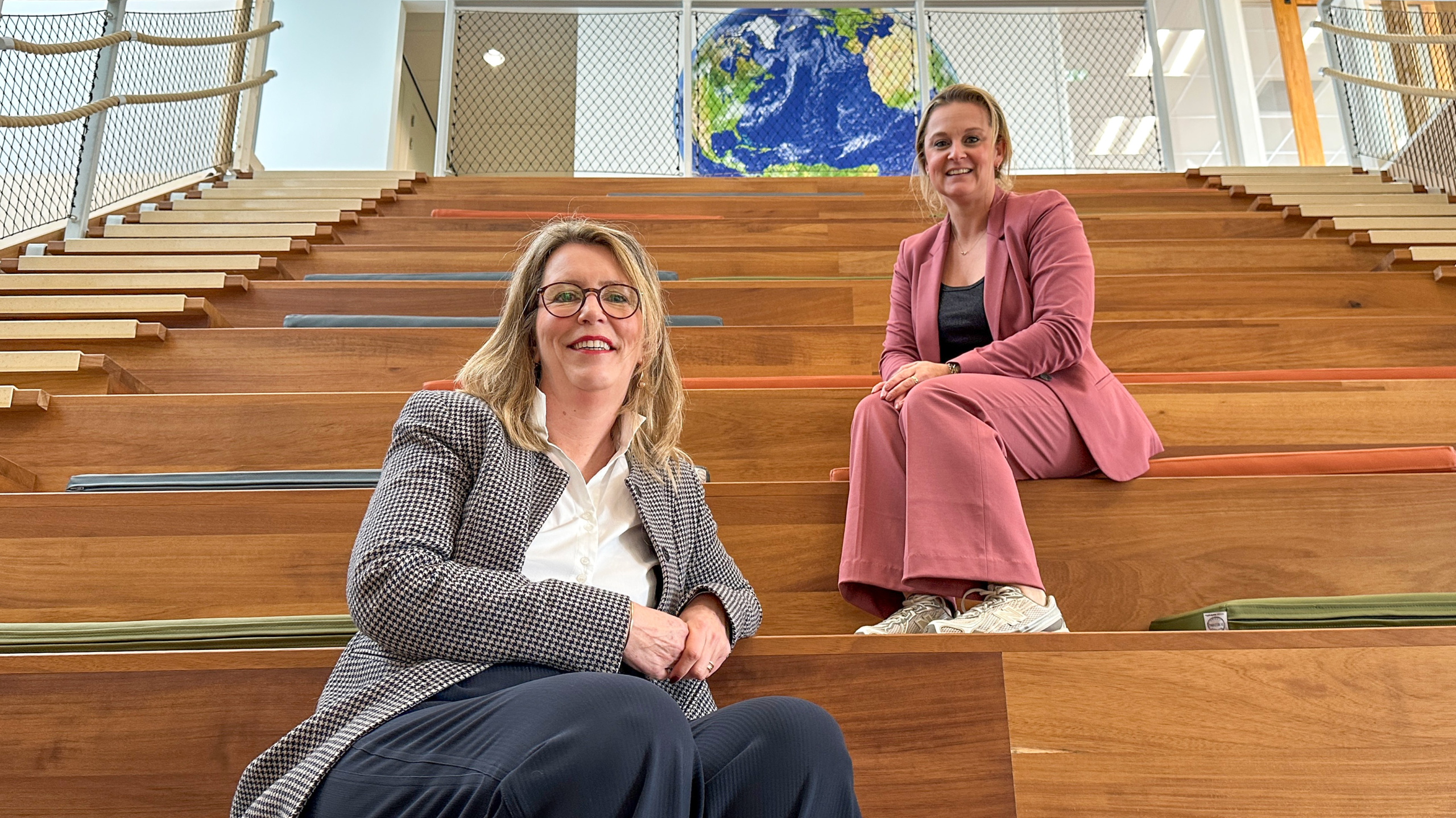 Twee vrouwen zitten op een houten trap met kleurrijke kussens in een moderne binnenruimte; op de achtergrond is een afbeelding van de aarde zichtbaar.