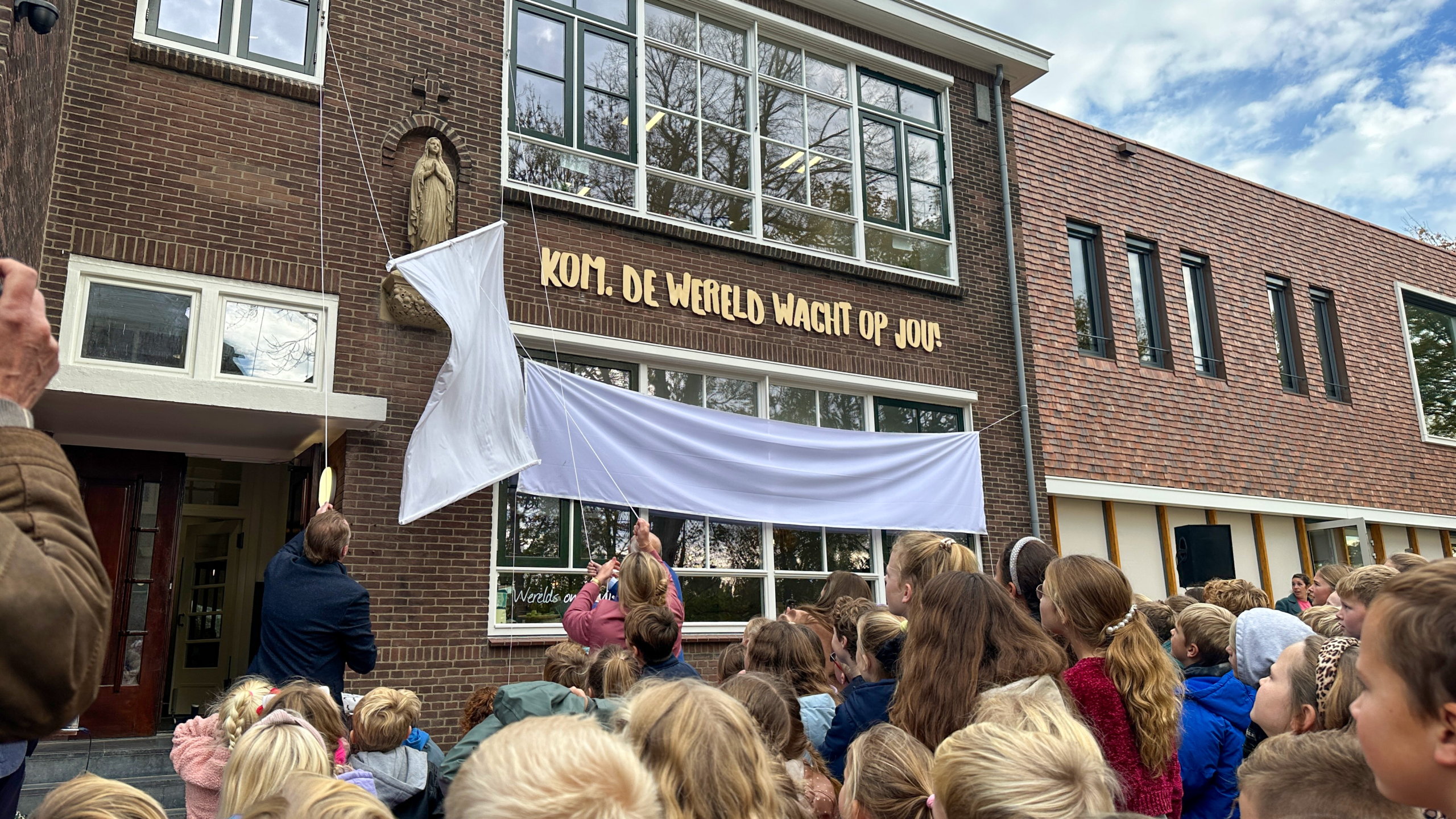 Groep kinderen verzameld voor een bakstenen gebouw, waar een doek wordt verwijderd om een tekst "Kom, de wereld wacht op jou!" te onthullen, met een beeld van een figuur erboven.