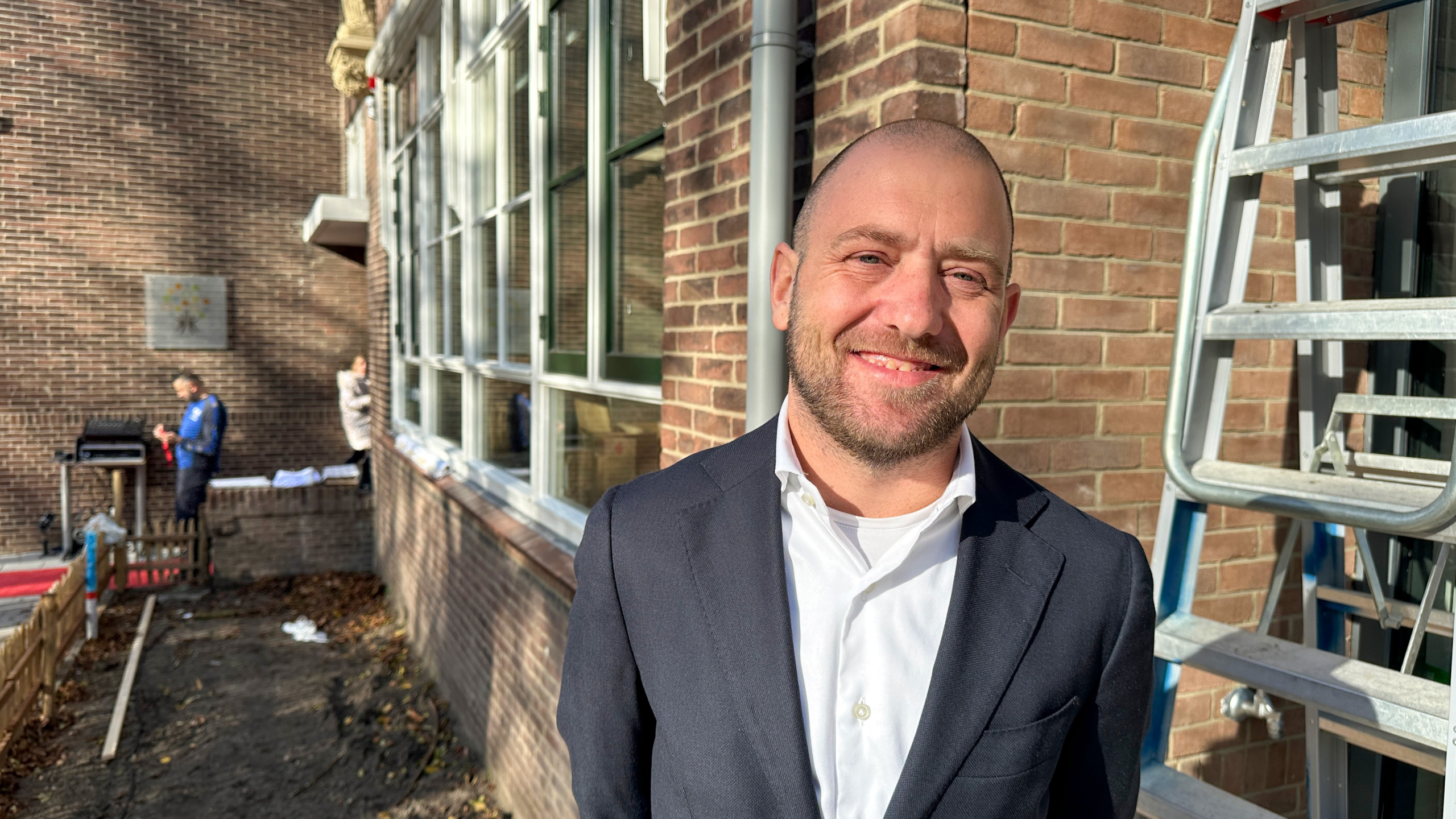 Een man in een donker pak en witte overhemd glimlacht naar de camera, staand naast een bakstenen muur met ramen, met zichtbare details van een ladder en twee mensen op de achtergrond in een buitenomgeving.