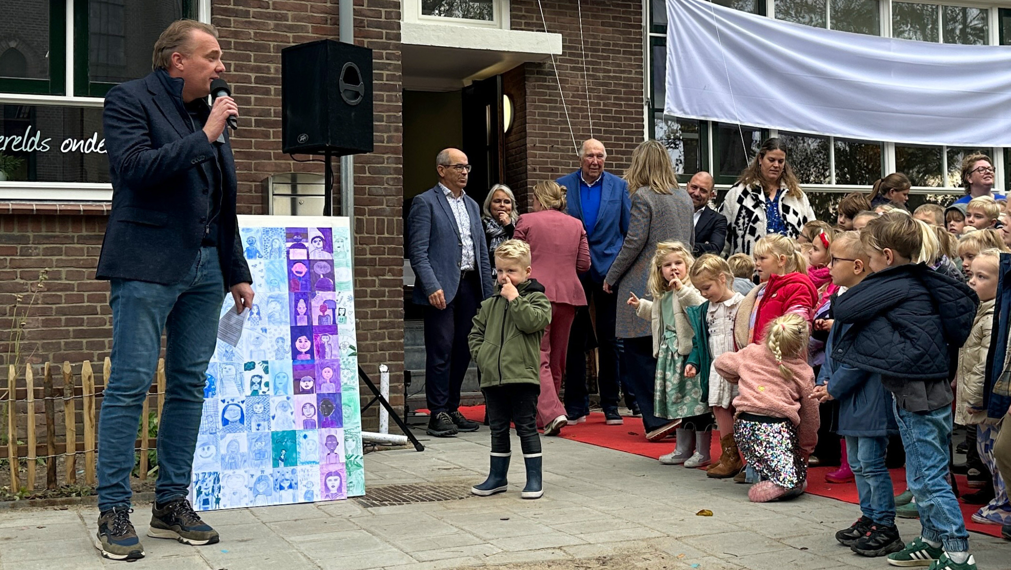 Een man spreekt in een microfoon buiten voor een gebouw met een groep kinderen en volwassenen. Op de achtergrond hangt een doek met de tekst 'Kom, de wereld wacht op jou!' op de muur. Kinderen staan op een rij voor het gebouw, sommige kijken naar de camera terwijl anderen naar de man luisteren.