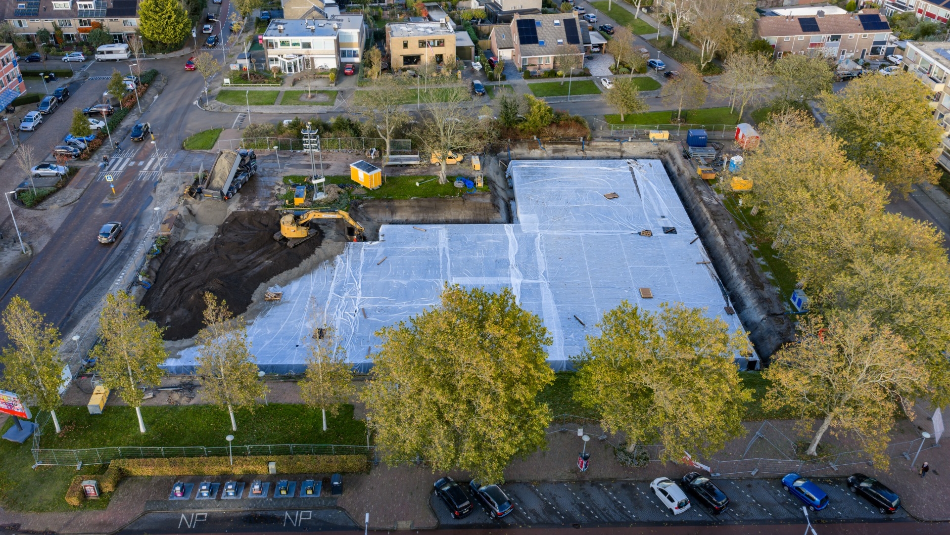 Luchtfoto van een bouwplaats in een stedelijke buurt, waarbij een groot deel is bedekt met witte zeilen. Een graafmachine is bezig met het verplaatsen van zand aan de linkerkant van de locatie. Er zijn bomen aan de rand van de bouwplaats en omringende woonhuizen.