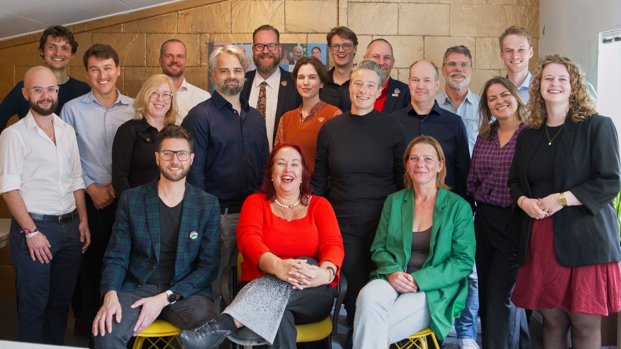 Groepsfoto van twintig mensen in een kantooromgeving. Sommigen zitten en anderen staan, allemaal glimlachen naar de camera. Diverse achtergrond en kledingstijlen, met een goudkleurige muur op de achtergrond.