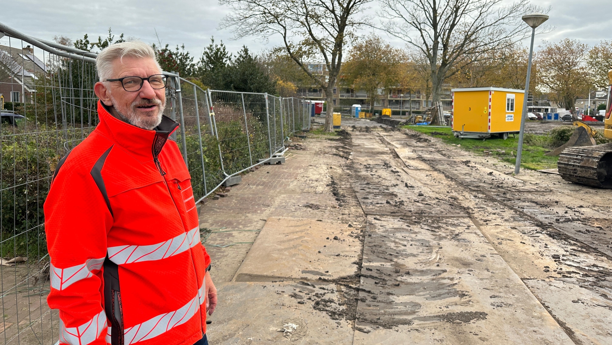 Man in a bright orange safety jacket standing at a construction site with a dirt path, surrounded by metal fencing and trees; a yellow portable cabin and construction equipment are visible in the background.
