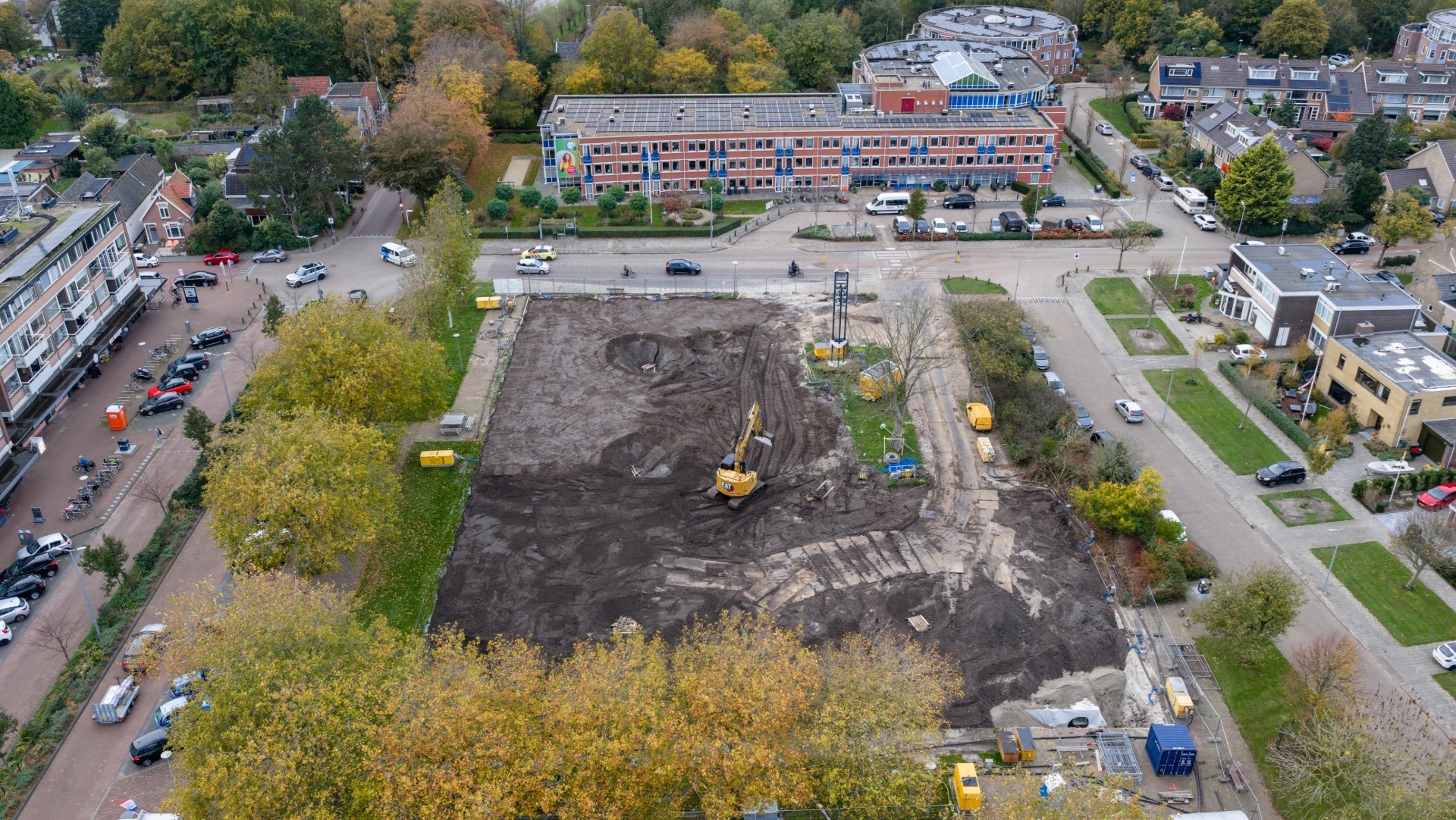 Luchtfoto van een bouwplaats omgeven door bomen en gebouwen, met een graafmachine aan het werk op een braakliggend terrein en verschillende geparkeerde voertuigen eromheen.
