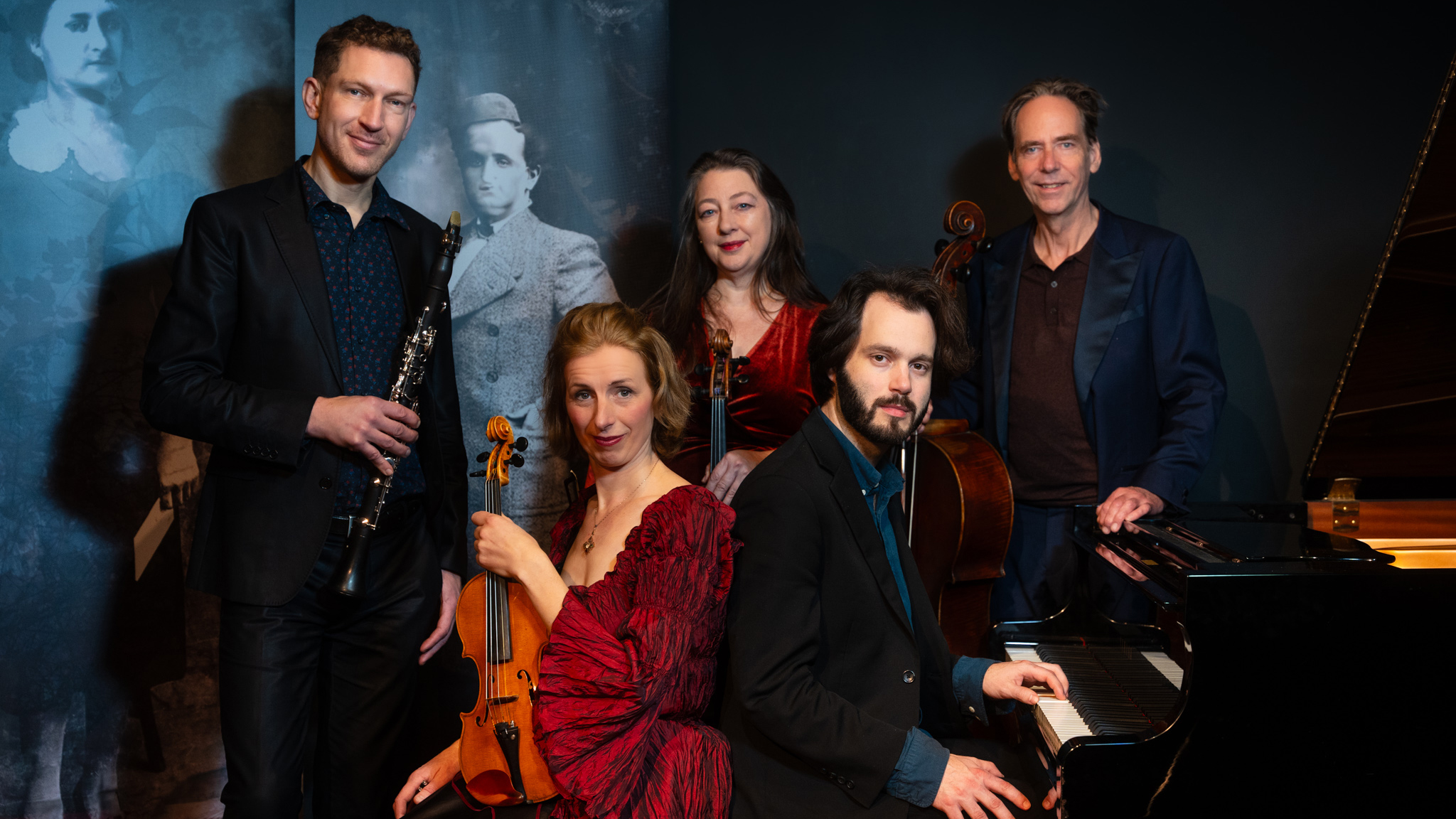 Een muziekensemble bestaande uit vijf personen poseert met hun instrumenten voor een donkere achtergrond, waaronder een piano, een klarinet, een viool en een cello.