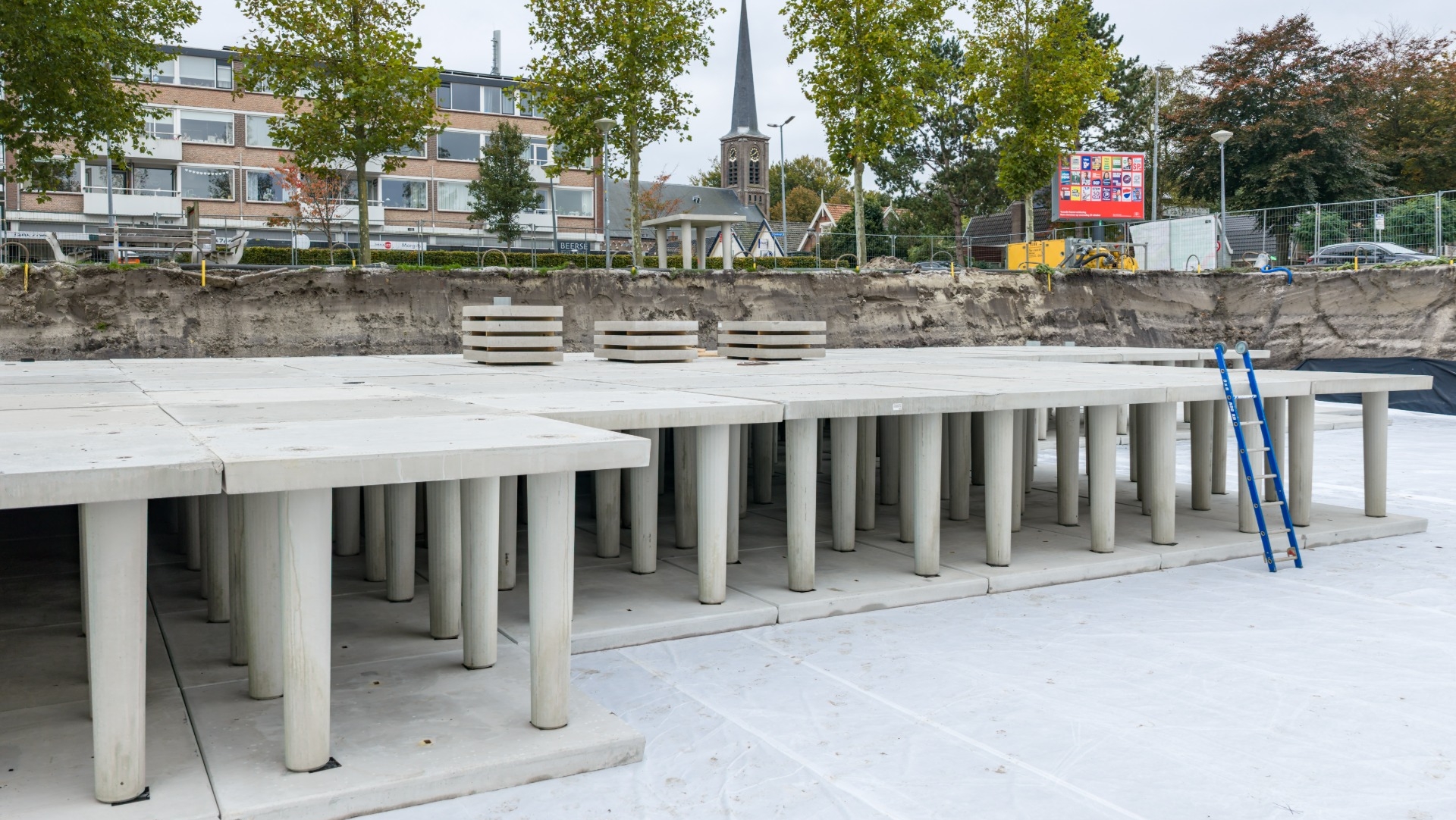 Betonnen constructie met pilaren in de bouwfase, tegen de achtergrond van een rij bomen en een kerk met een torenspits. Op de achtergrond zijn een gebouw en een hek zichtbaar. Er staat een blauw laddertje tegen de constructie aan.