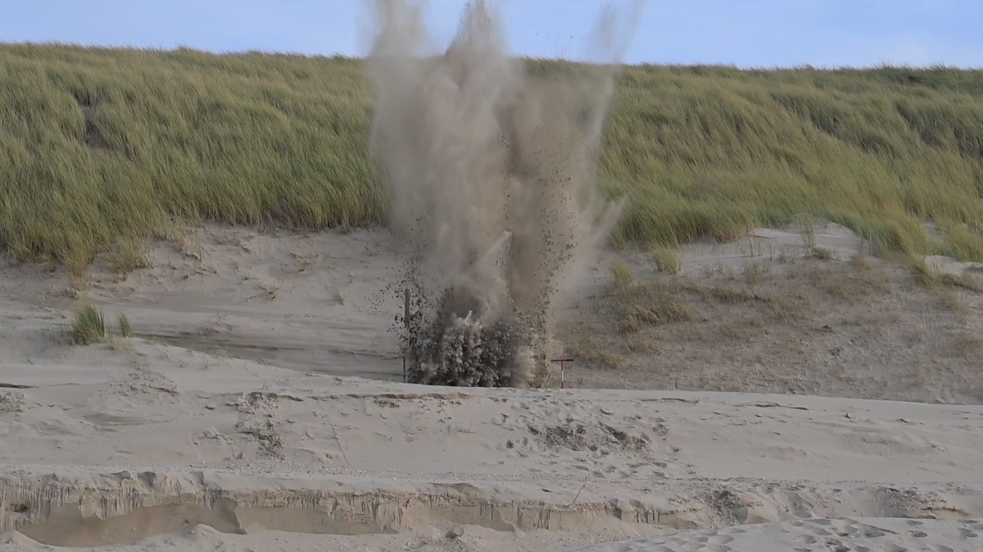 Een explosie van zand en stof in een duinlandschap met gras op de achtergrond.