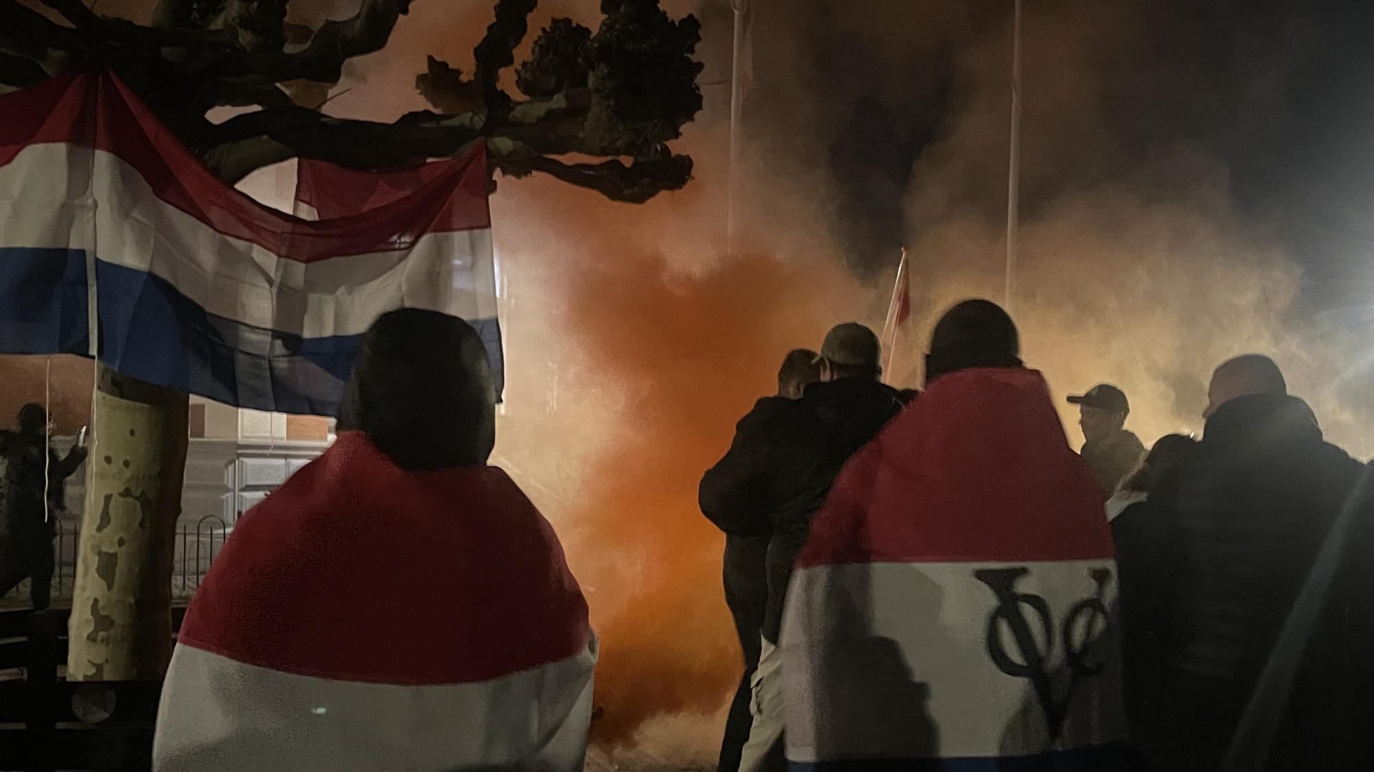 Een groep mensen verzamelt zich 's nachts met rook en vlaggen, en sommigen zijn gehuld in wat lijkt op Nederlandse vlaggen.