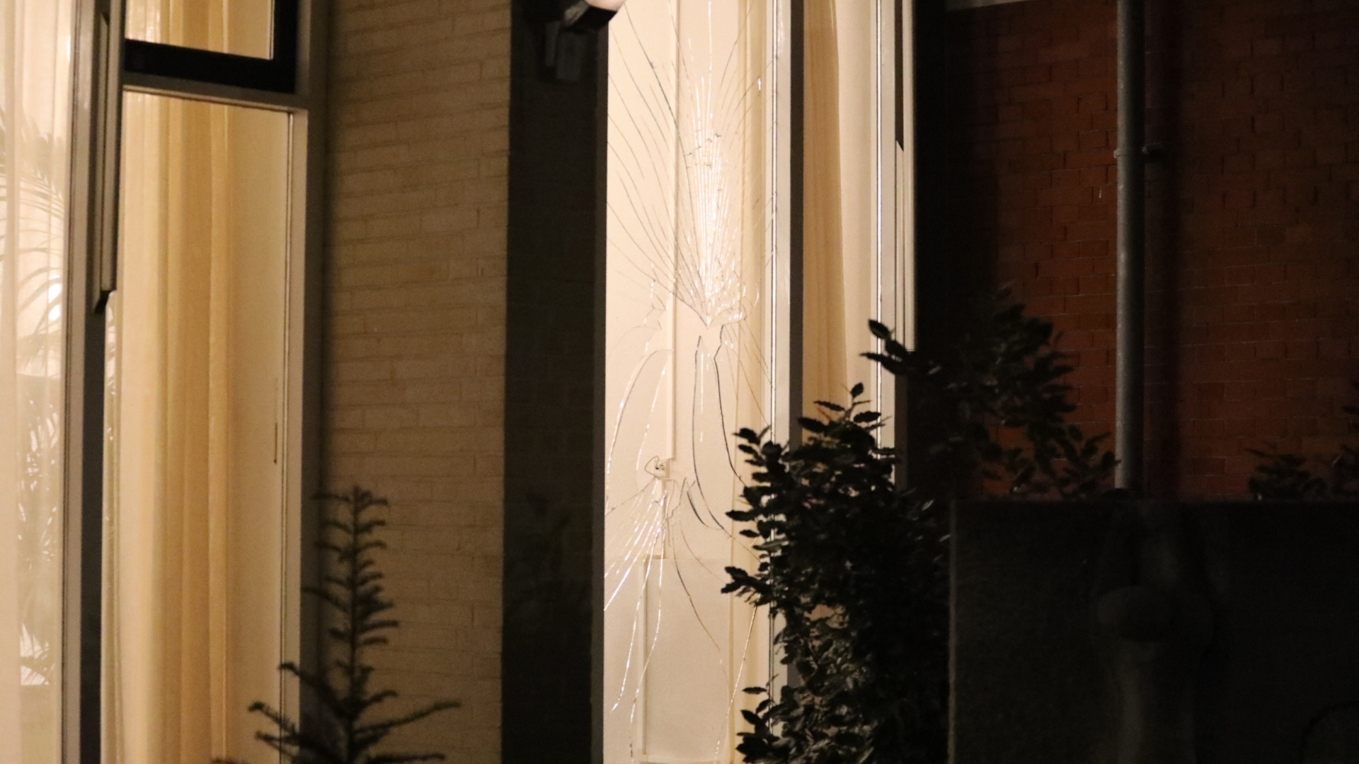 Nachtelijke buitenaanzicht van een raam met gebroken glas door een impact; er is licht van binnen zichtbaar en omringende struiken zijn in de schaduw te zien.