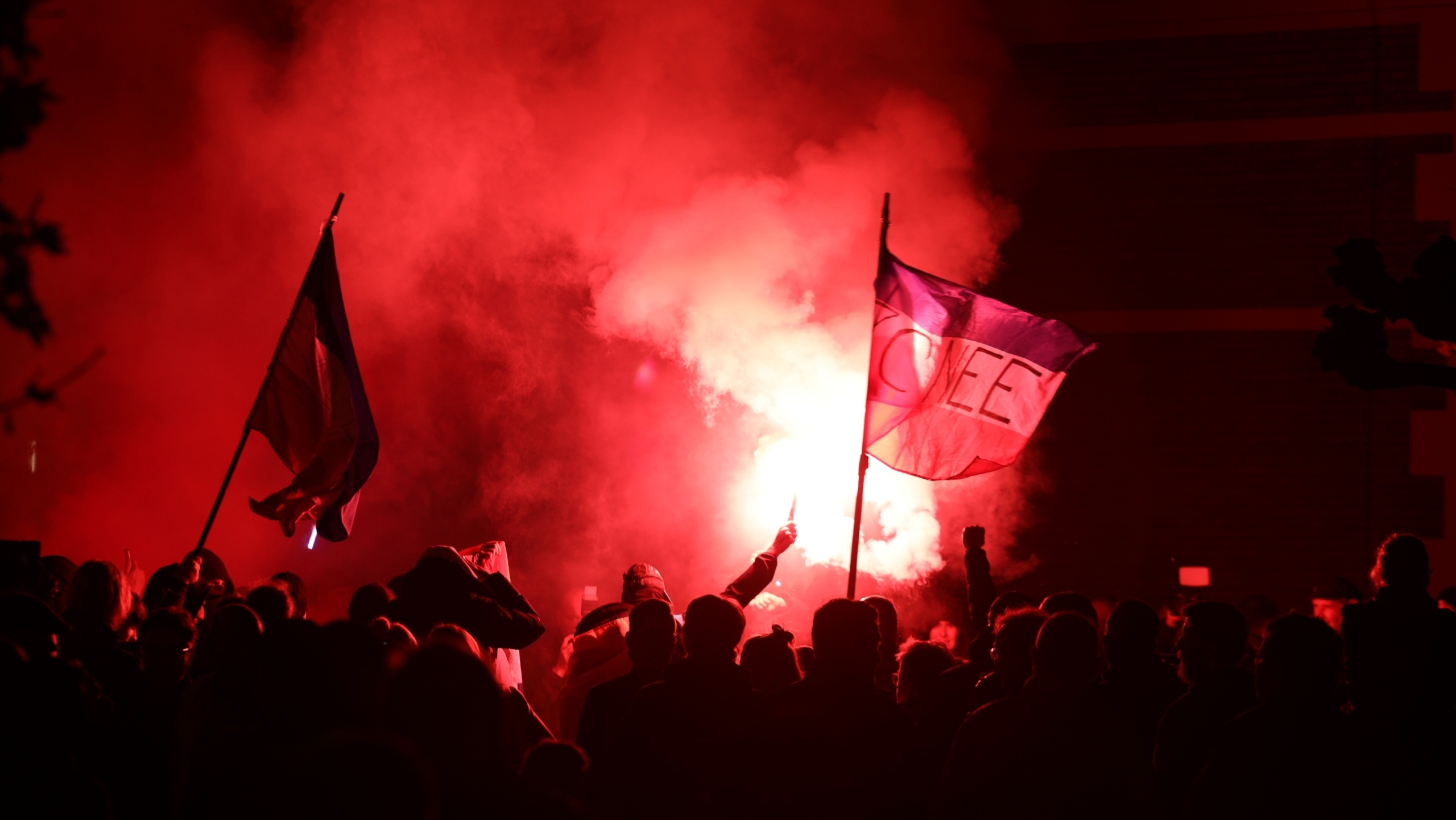 Een menigte mensen met vlaggen en fakkels staat in het donker, verlicht door een felrode gloed van vuurwerk.