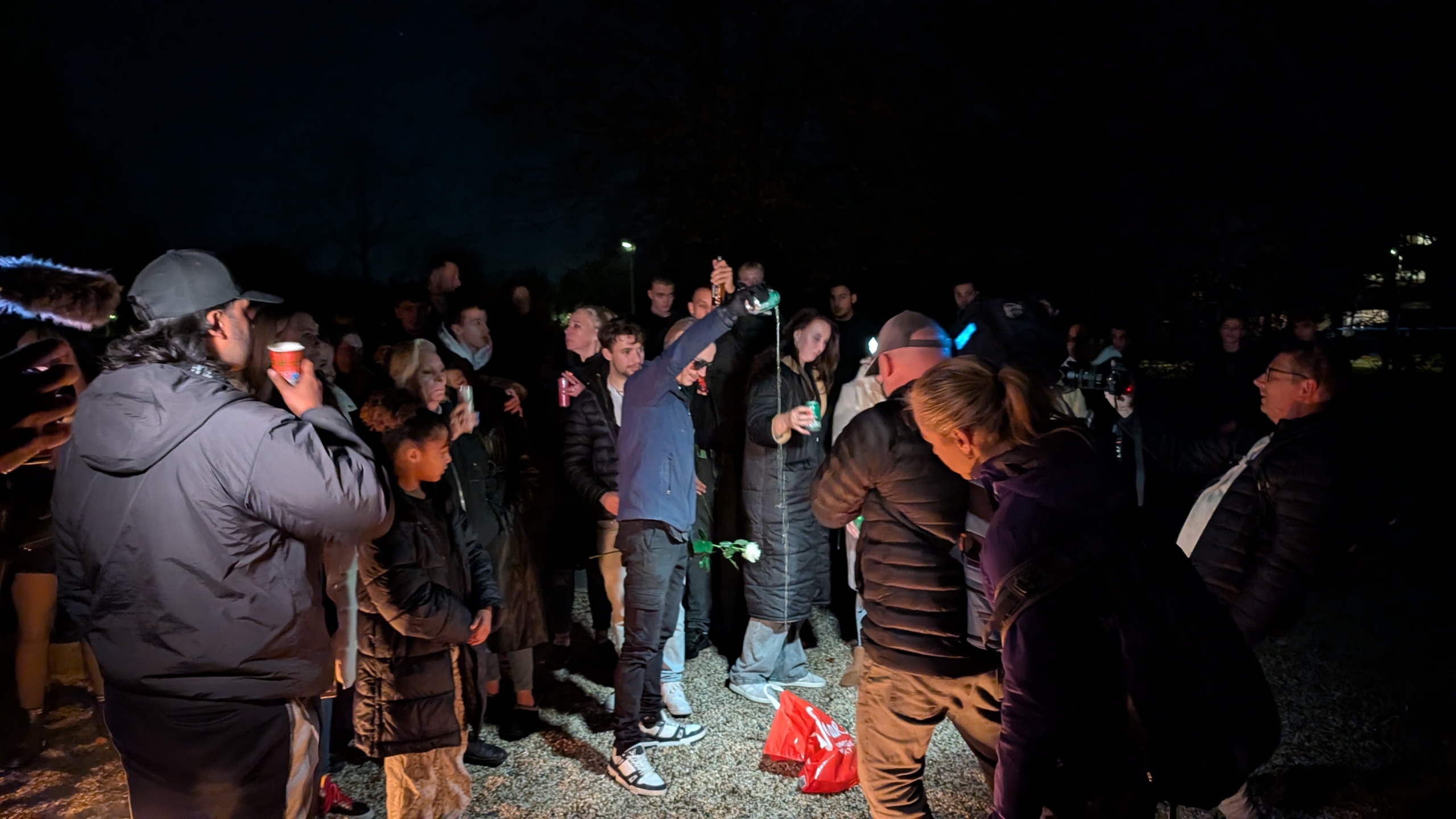Groep mensen staat in het donker bijeen, sommigen met drankjes in de hand, terwijl een persoon in het midden een fles omhooghoudt waar vloeistof uit stroomt.