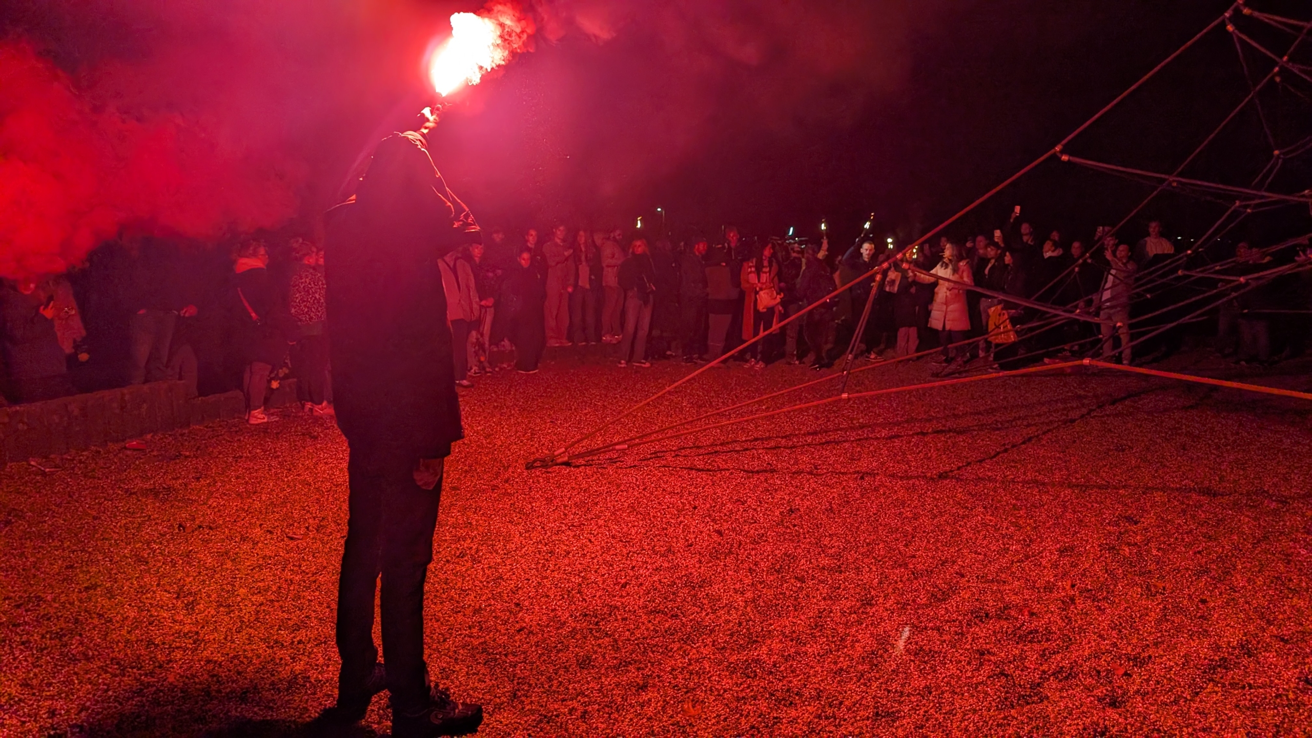 Iemand houdt een fakkel vast die fel rood licht uitstraalt, met een menigte van mensen die vanaf de zijkant toekijkt, 's nachts in een open ruimte.