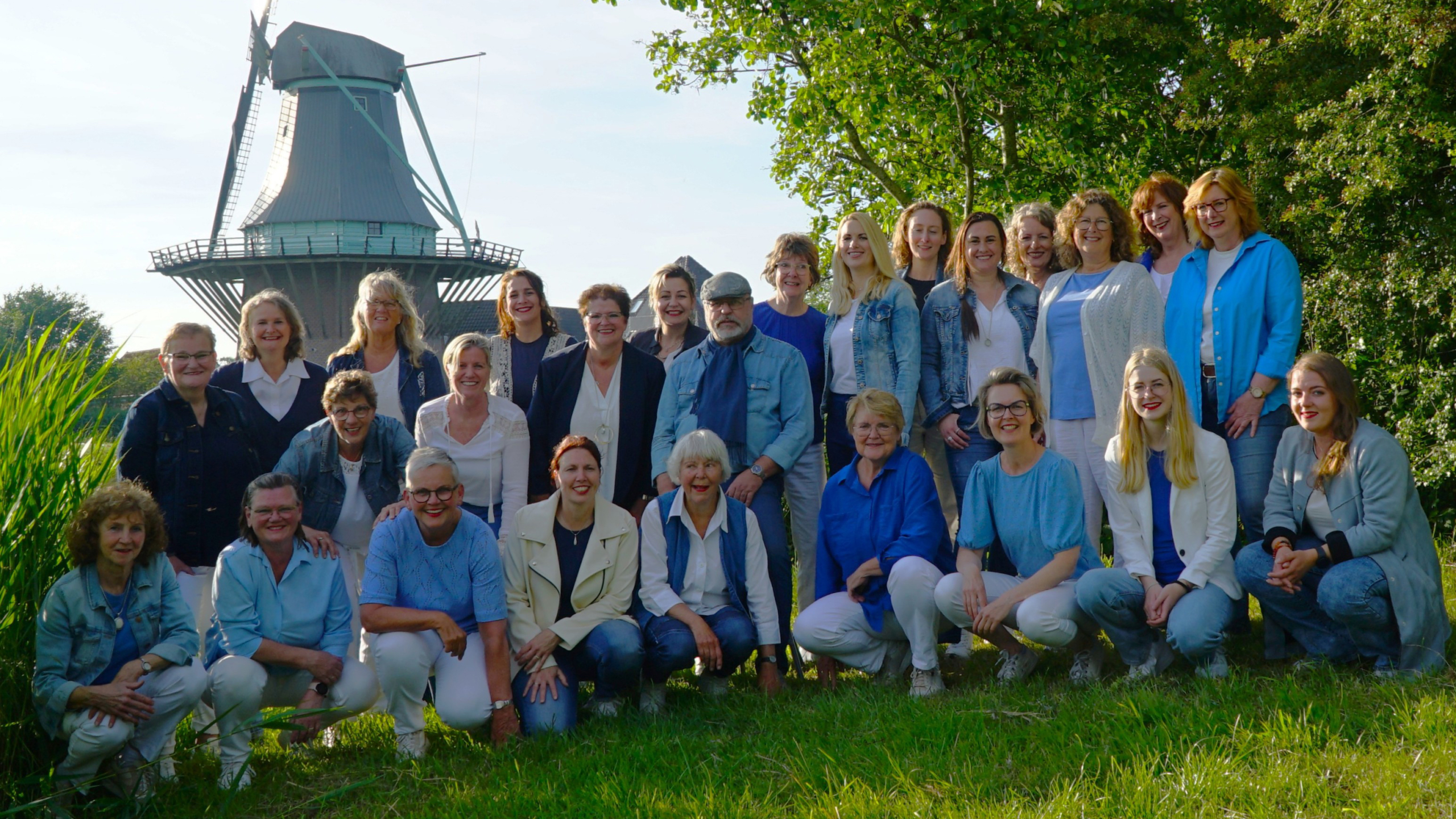Een groep mensen poseert in het gras voor een traditionele Nederlandse windmolen, omringd door groen en een heldere lucht.