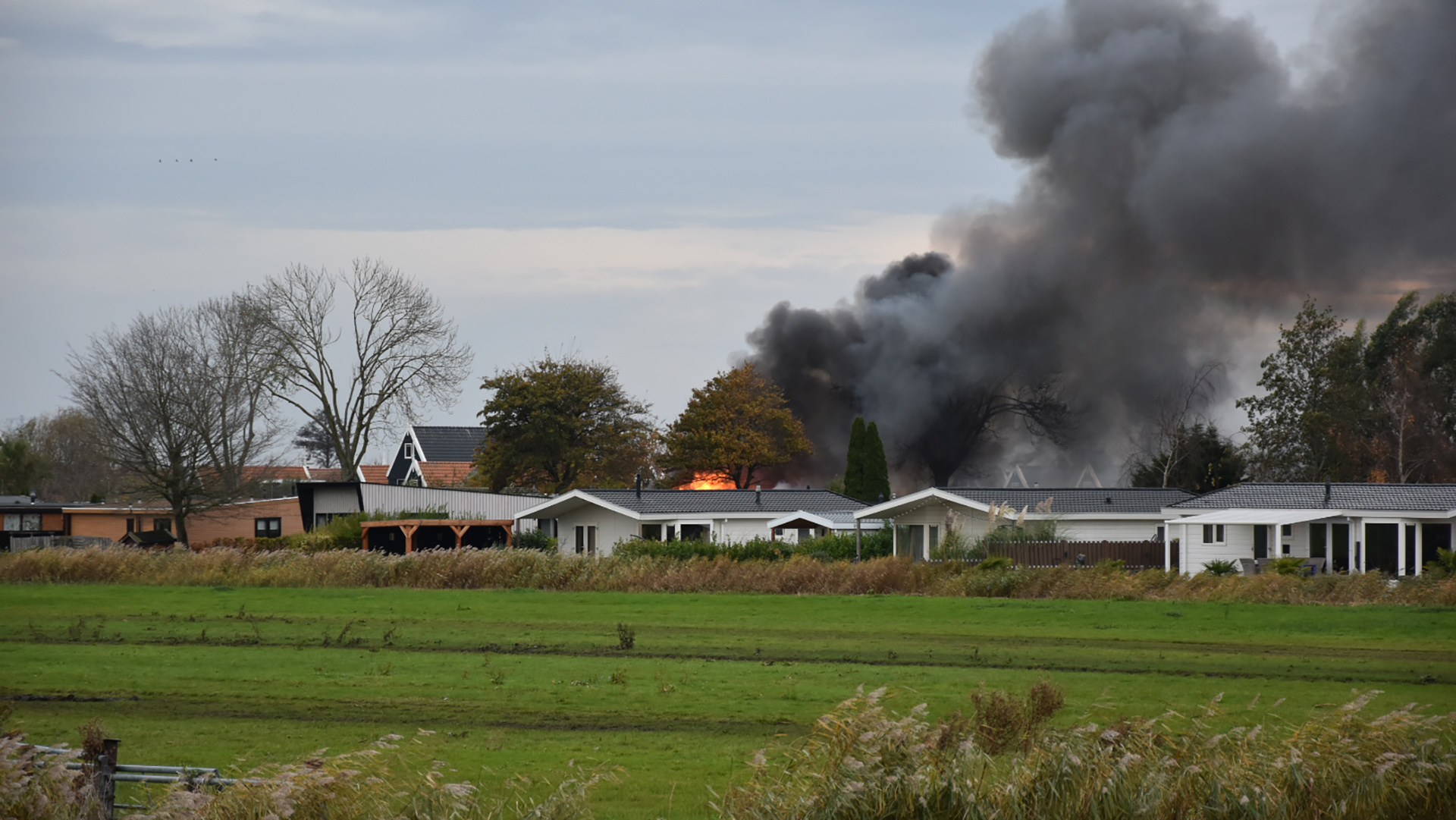 Een vuurbrand met dikke zwarte rook stijgt op achter huizen in een groene landelijke omgeving.