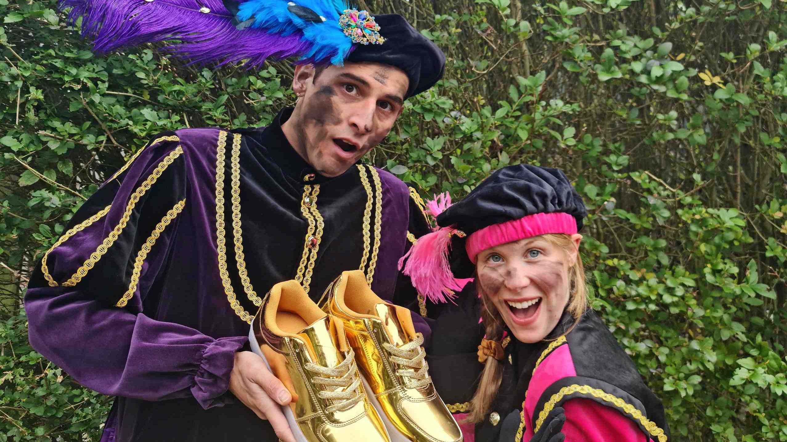 Twee mensen verkleed als Zwarte Pieten poseren buiten met gouden sportschoenen in hun handen.