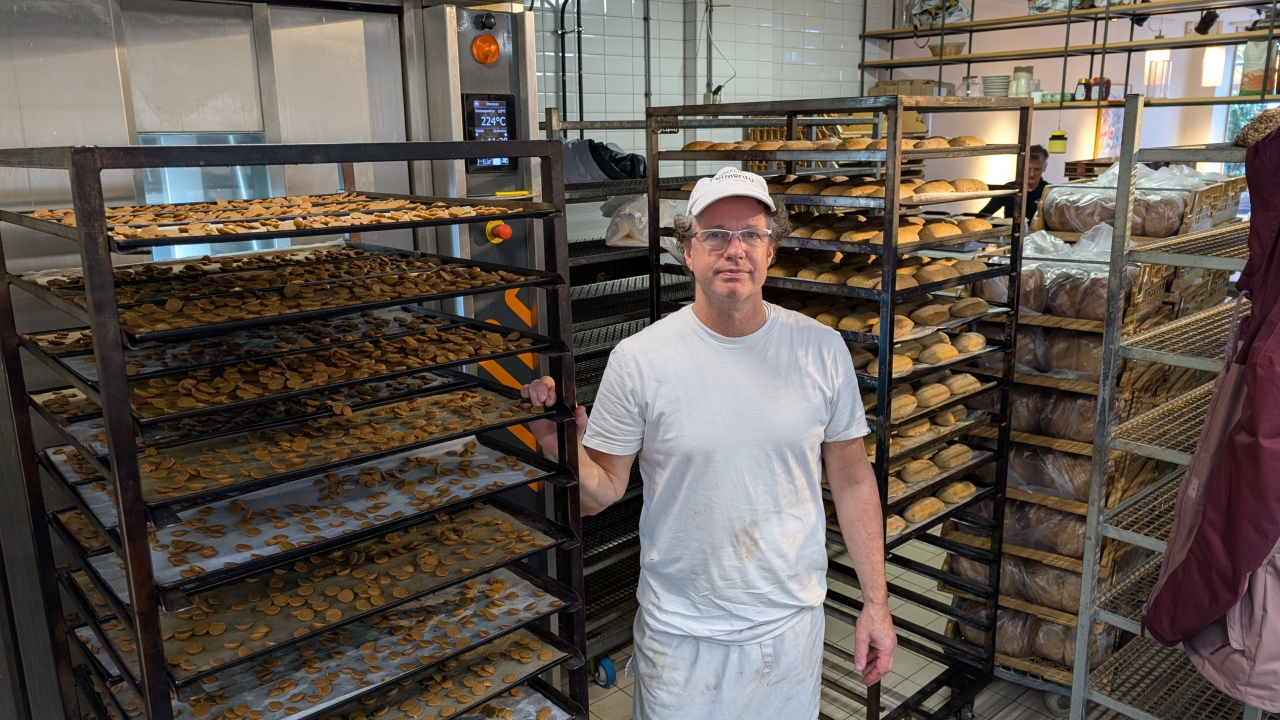 Een bakker in witte kleding staat in een bakkerij naast rekken vol met koekjes en brood.