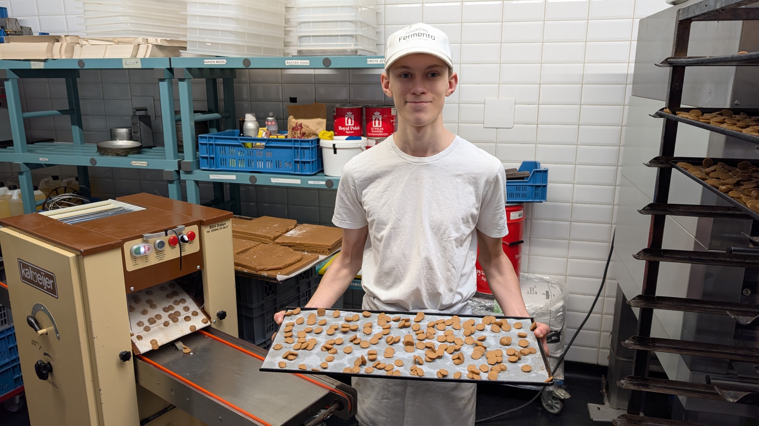 Jonge man in een bakkerij, gekleed in een wit T-shirt en een witte pet, houdt een bakplaat met ongebakken koekjes vast. Op de achtergrond zijn een machine en bakkerijbenodigdheden te zien.