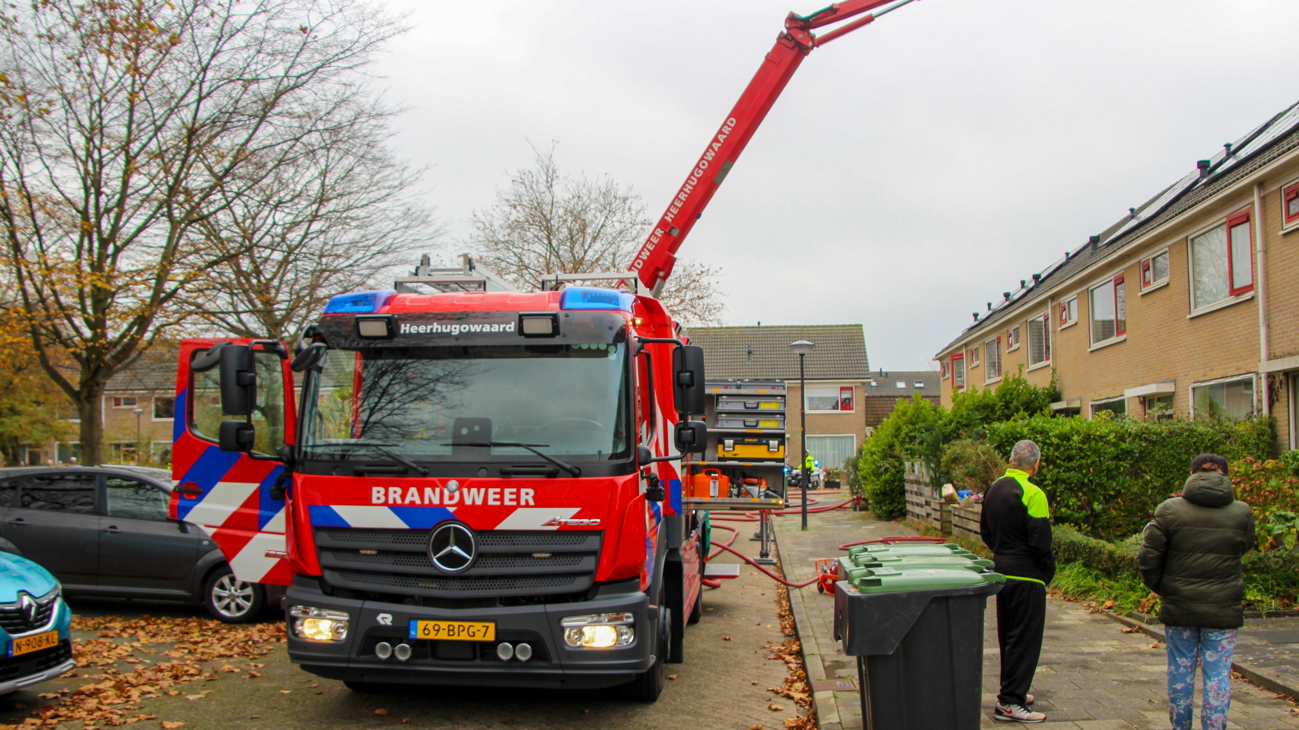 Brandweerwagen met opschrift 'Heerhugowaard' staat geparkeerd in een woonwijk, met een uitgeschoven kraan bij een rijtjeshuis. Twee mensen staan op de stoep te kijken.