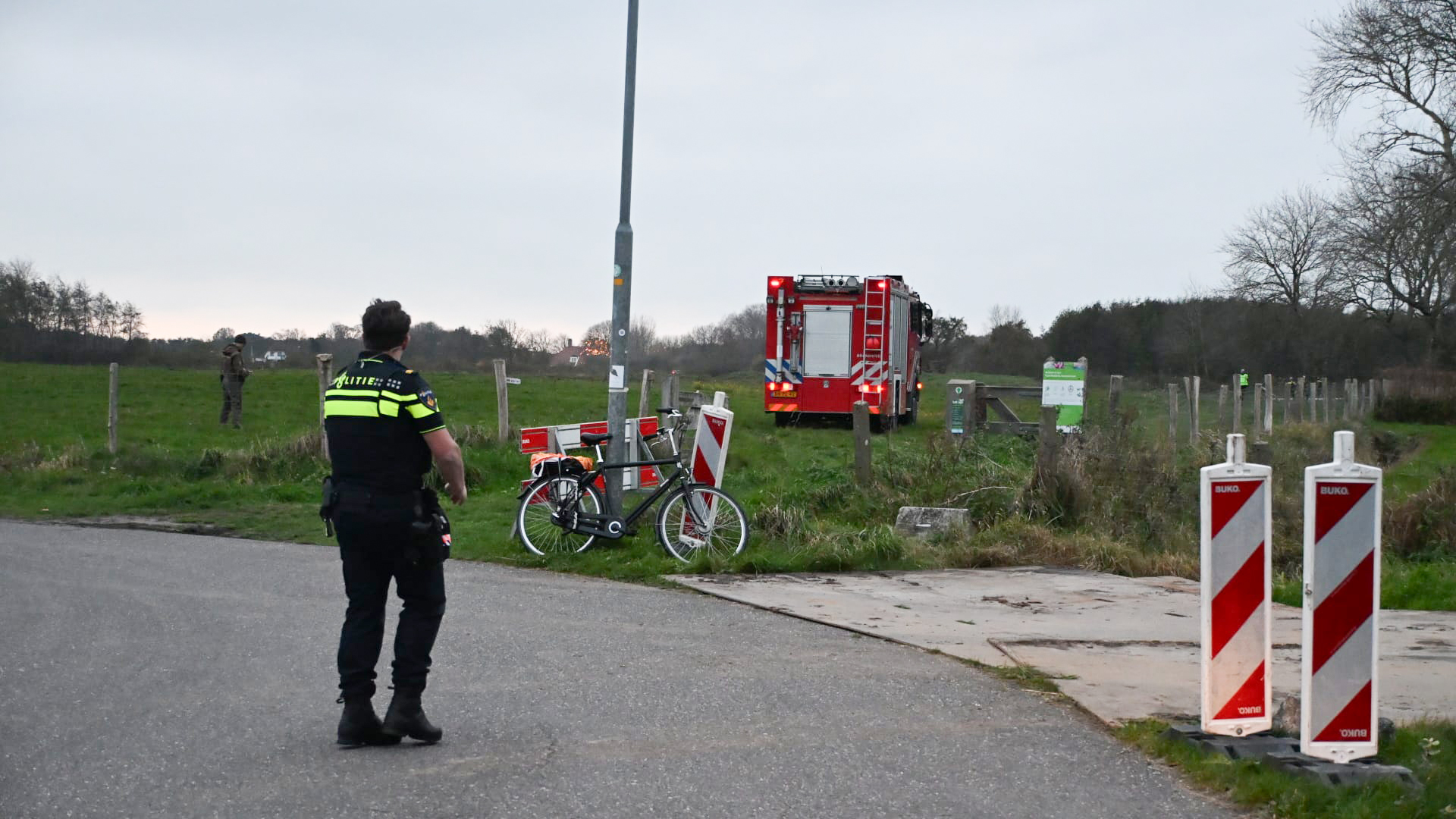 Politieagent bij een landweg kijkt in de richting van een brandweerwagen die op een grasveld staat geparkeerd, met een paar mensen aan de zijlijn; naast een lantaarnpaal staat een fiets.