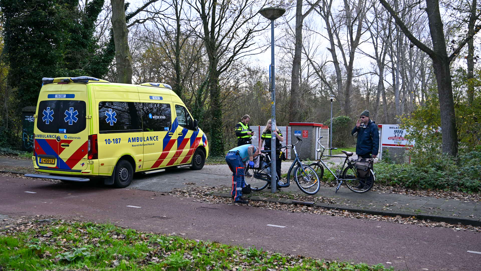 Een gele ambulance staat geparkeerd langs een pad naast een grasveld, terwijl twee politieagenten en een man bij een fiets staan. Een andere man met een fiets staat op de achtergrond. Er zijn bomen en bladeren te zien in de omgeving.