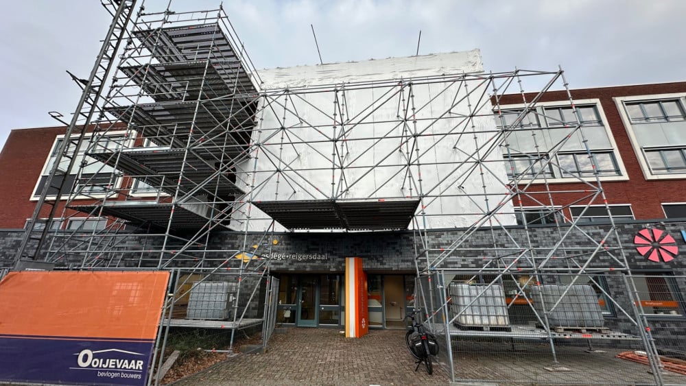 Gebouw in renovatie met steigers aan de gevel, omgeven door hekken en bouwmaterialen, met een entree voorzien van oranje en gele decoraties.
