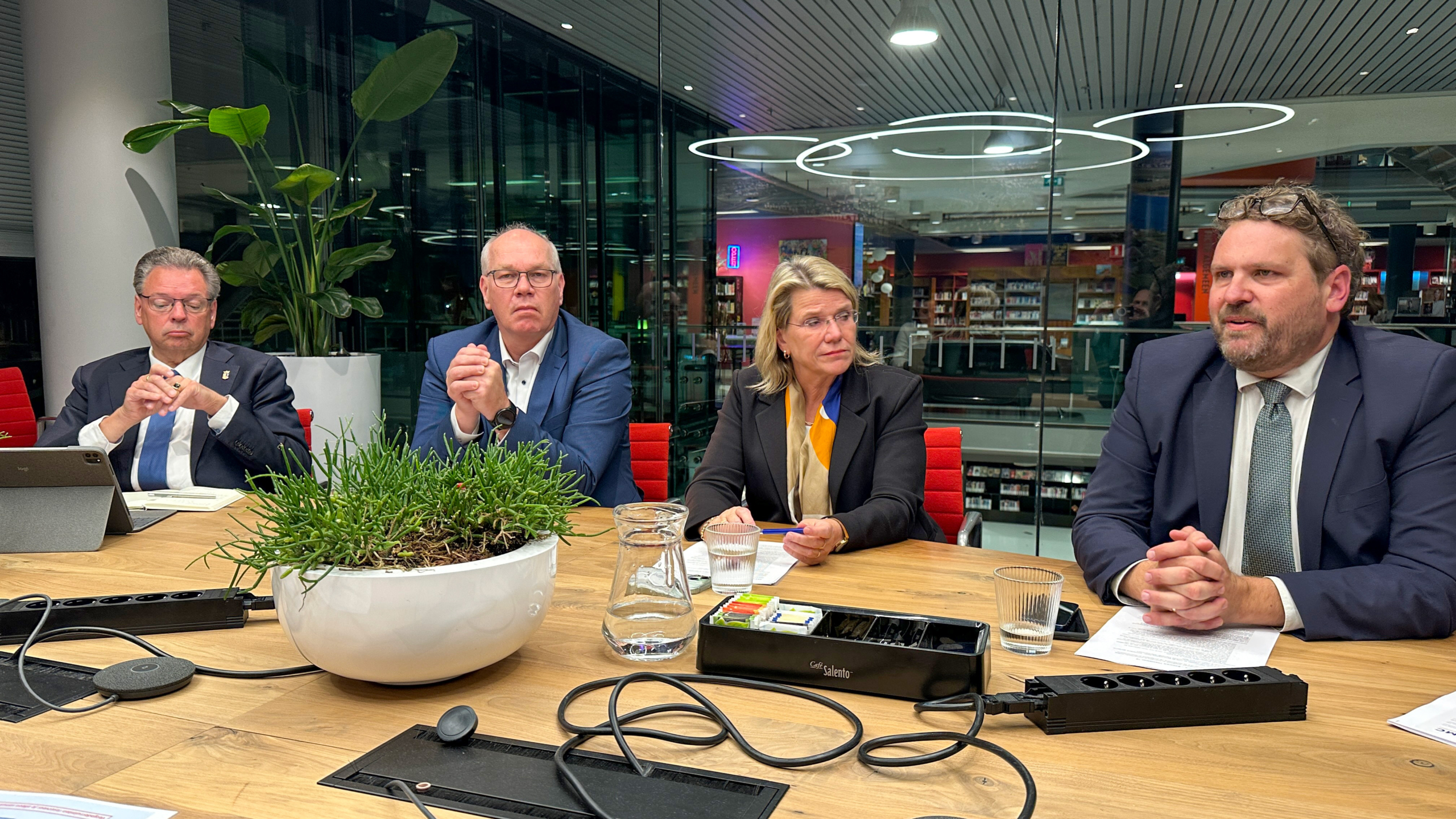 Vier personen in formele kleding zitten aan een tafel in een moderne vergaderruimte met glazen wanden en een schaal met planten op de tafel.