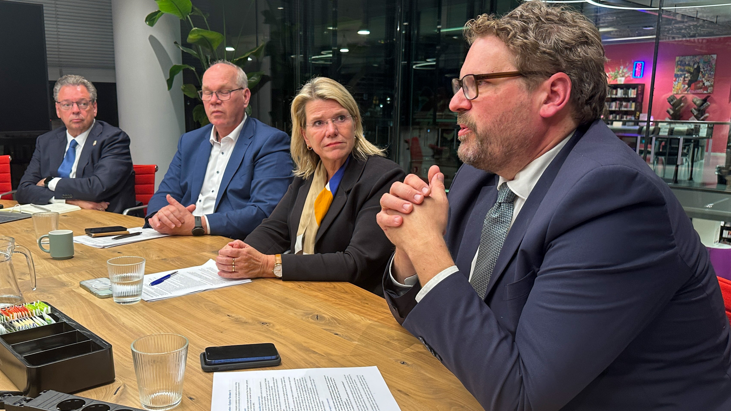 Vier mensen in zakelijke kleding zitten aan een tafel tijdens een vergadering, met notities en glazen water voor hen.