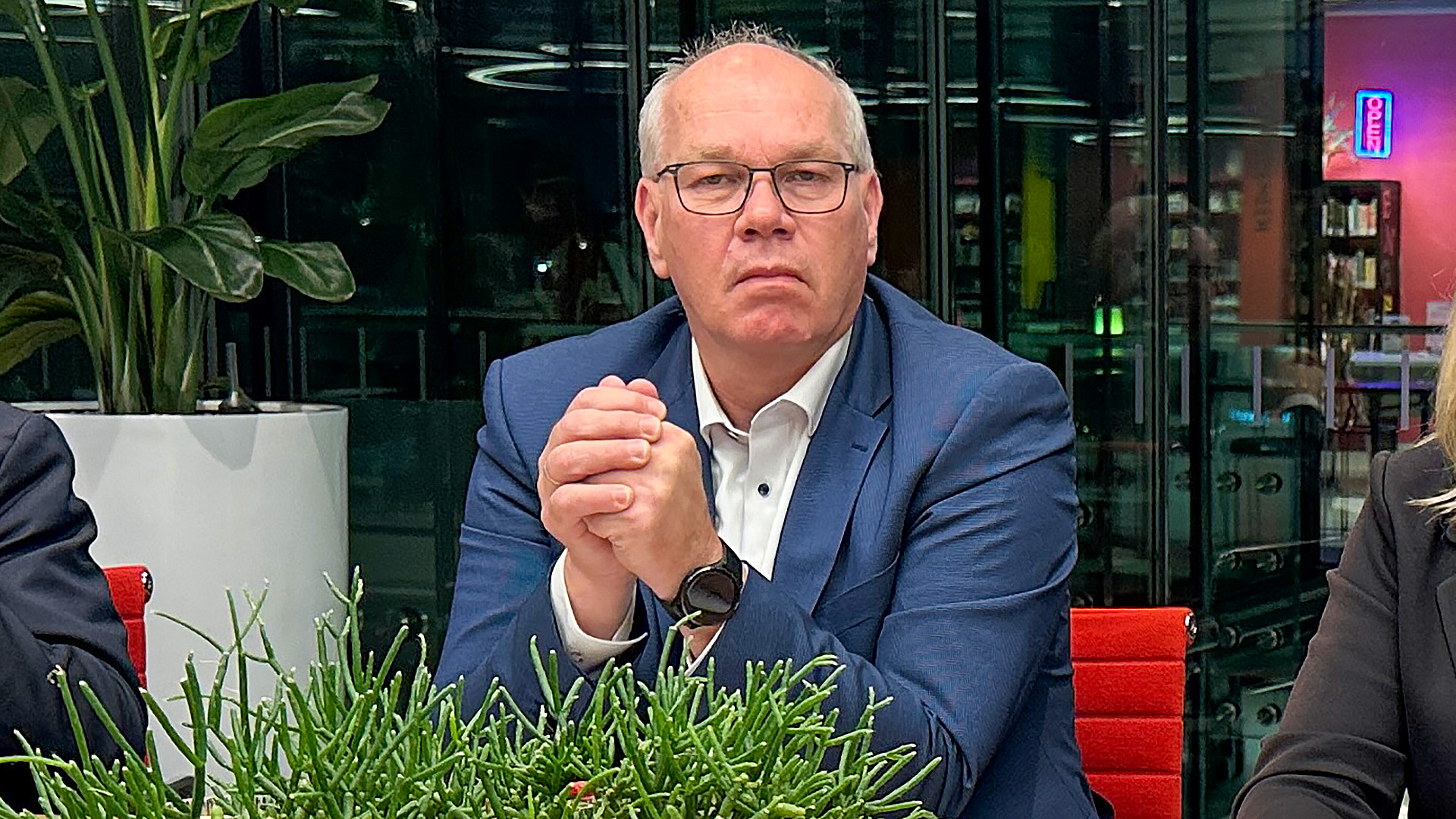 Een man met een bril en een blauw pak zit aan een tafel met gevouwen handen, omringd door planten en een moderne, verlichte achtergrond.