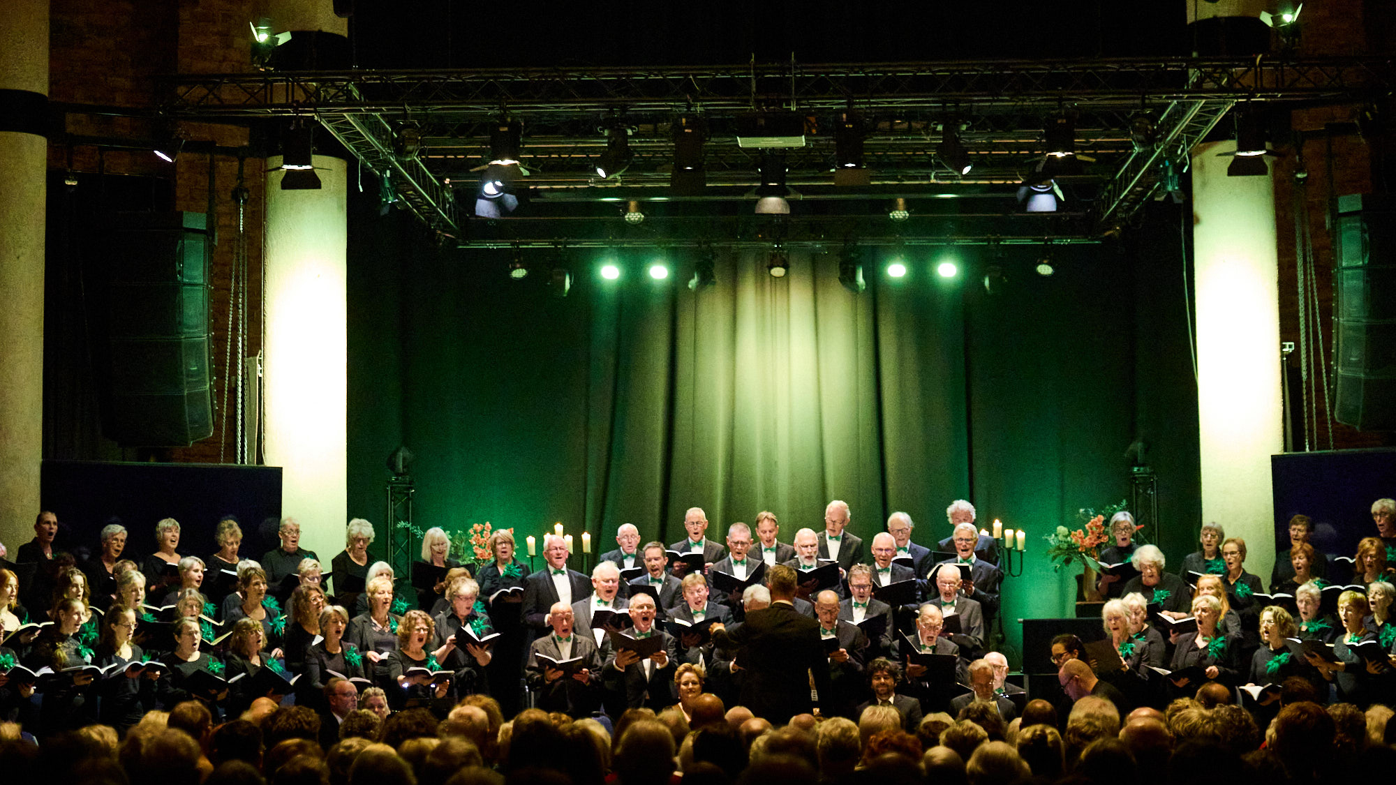 Groot koor in zwarte kleding zingt op een verlicht podium met groene achtergrondverlichting.