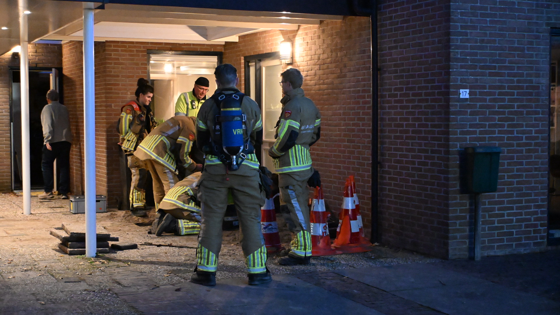 Brandweerlieden staan voor een gebouw en voeren een taak uit, met verschillende veiligheidskegels op de grond.