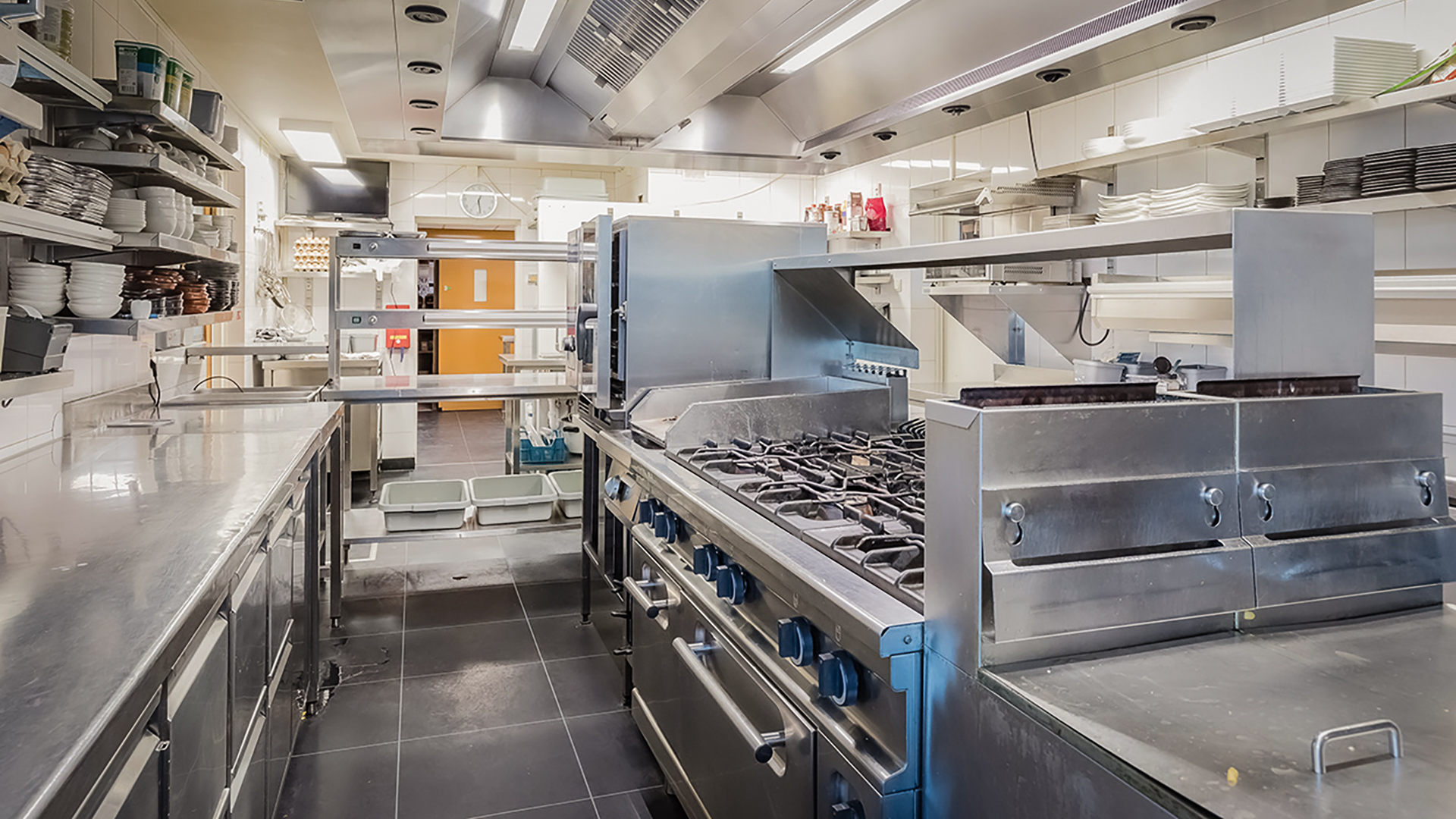 Een industriële keuken met roestvrijstalen apparatuur, waaronder een groot fornuis met meerdere branders, omringd door arbeidsoppervlakken en planken vol borden en keukenbenodigdheden.