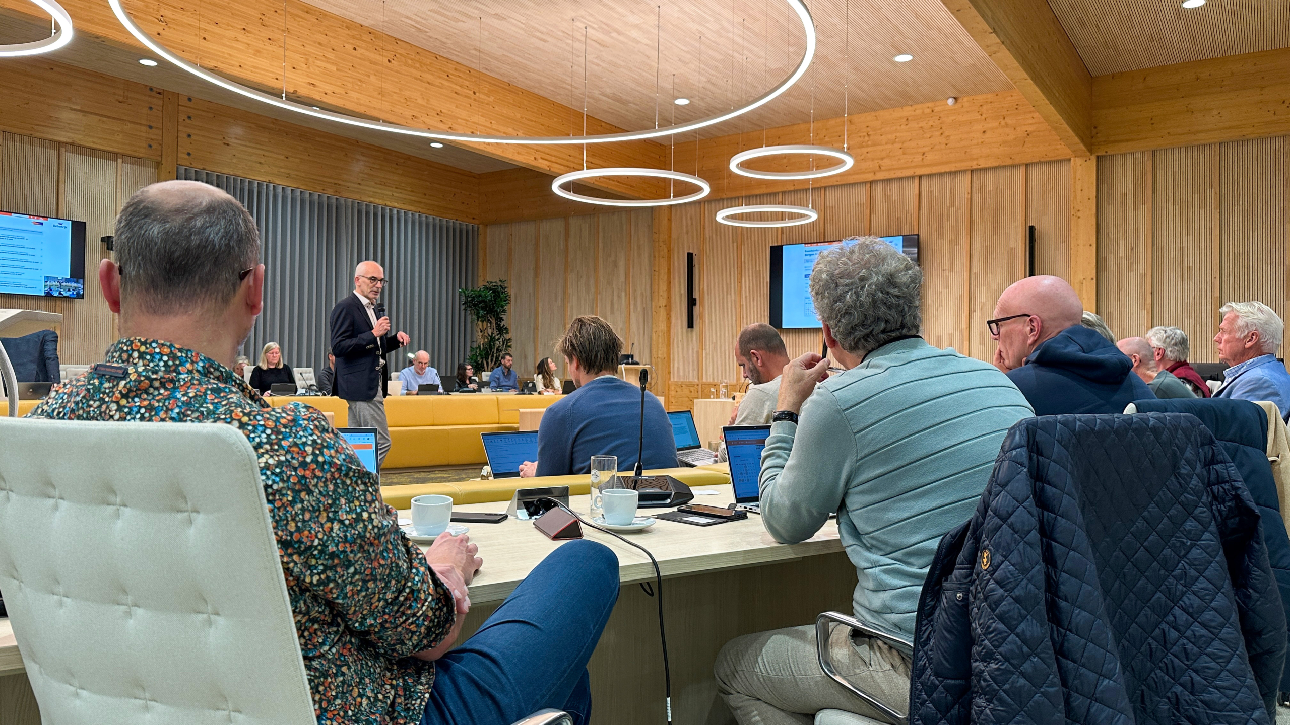 Groep mensen in een vergaderruimte met houten muren, zittend rond een tafel met laptops en papieren, terwijl een spreker met microfoon aan het presenteren is.