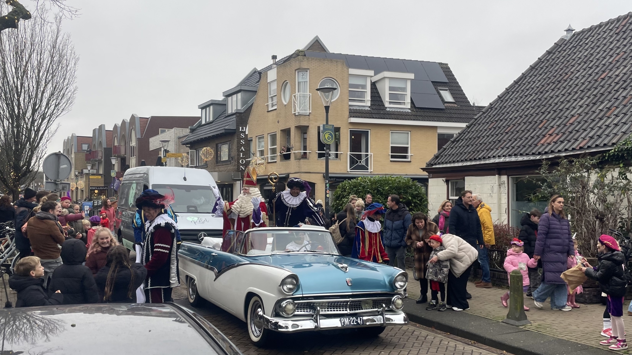 Een optocht met een klassieke auto en feestelijk verklede mensen in een straat vol toeschouwers.