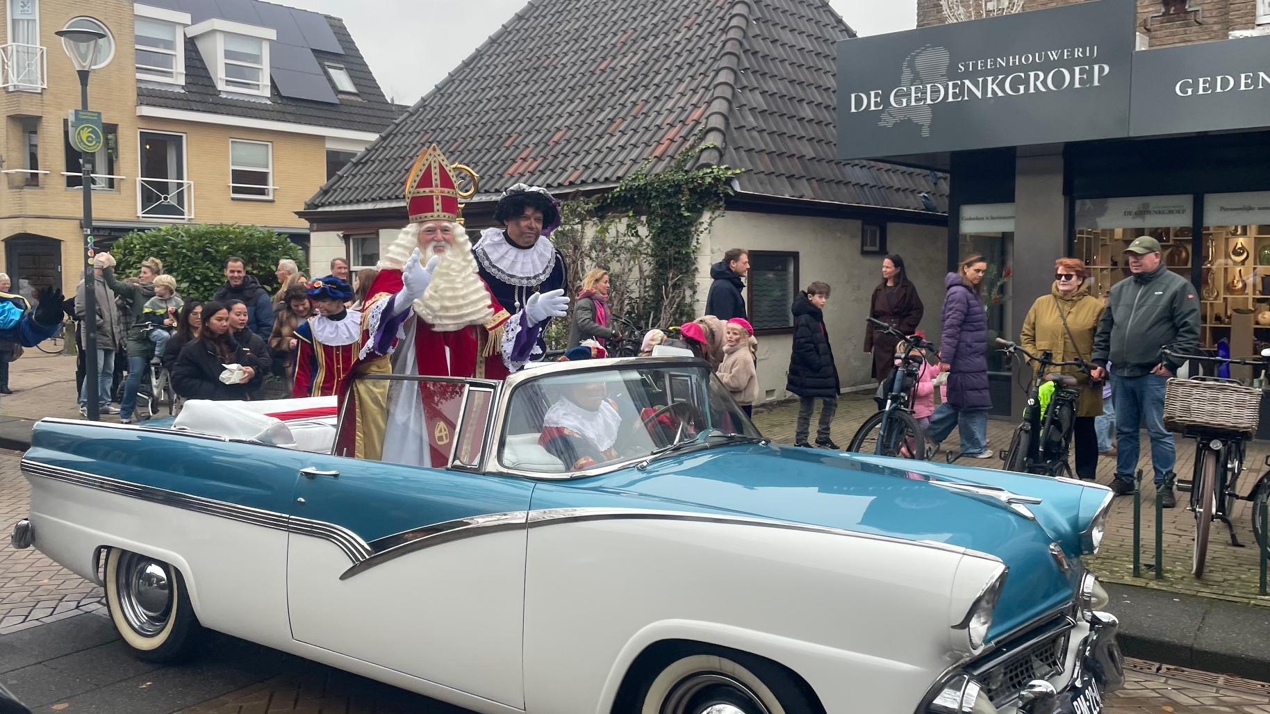 Sinterklaas en Pieten rijden in een klassieke blauwe en witte cabriolet langs een straat met toeschouwers, tijdens een Sinterklaasintocht.