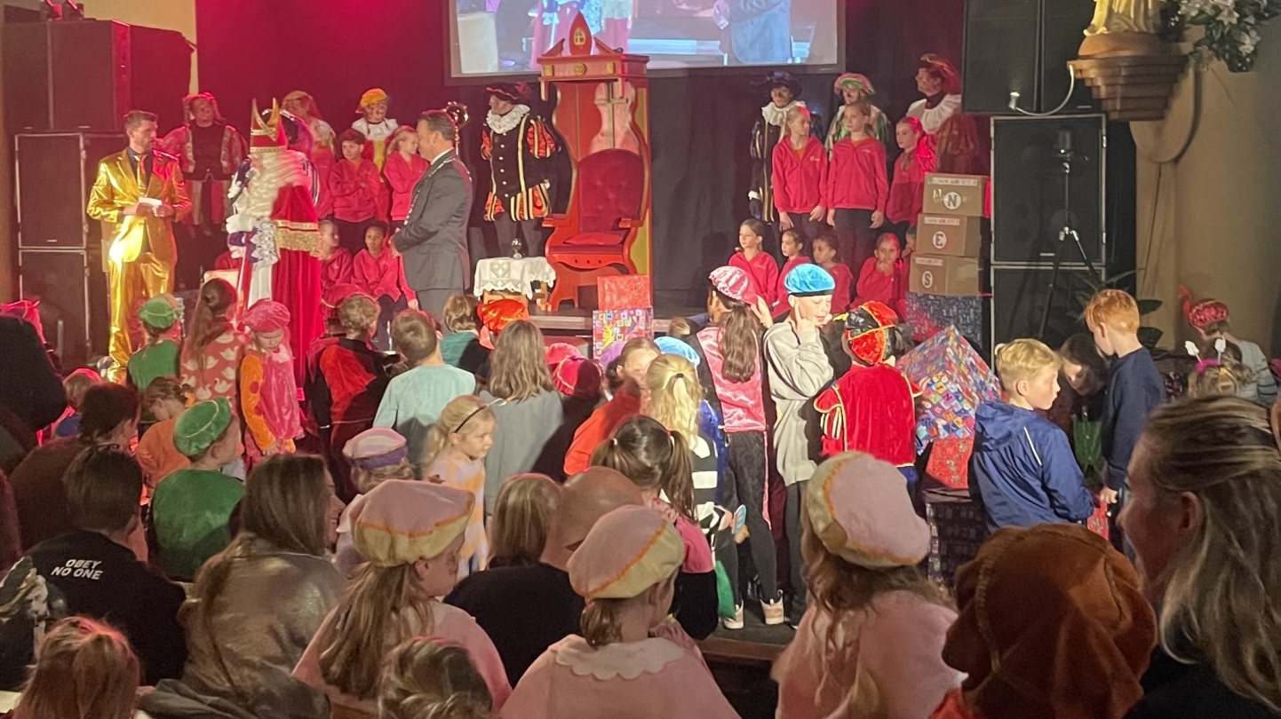Een Sinterklaasviering in een kerk, met een menigte kinderen en volwassenen, veelal in kostuums, die naar het podium kijken waar een Sinterklaasfiguur staat. De omringende kinderen dragen kleurrijke pietenkostuums en mutsen. Boven het podium hangt een groot scherm dat het gebeuren toont. De omgeving heeft gotische bogen en beelden, passend bij een kerkinterieur.