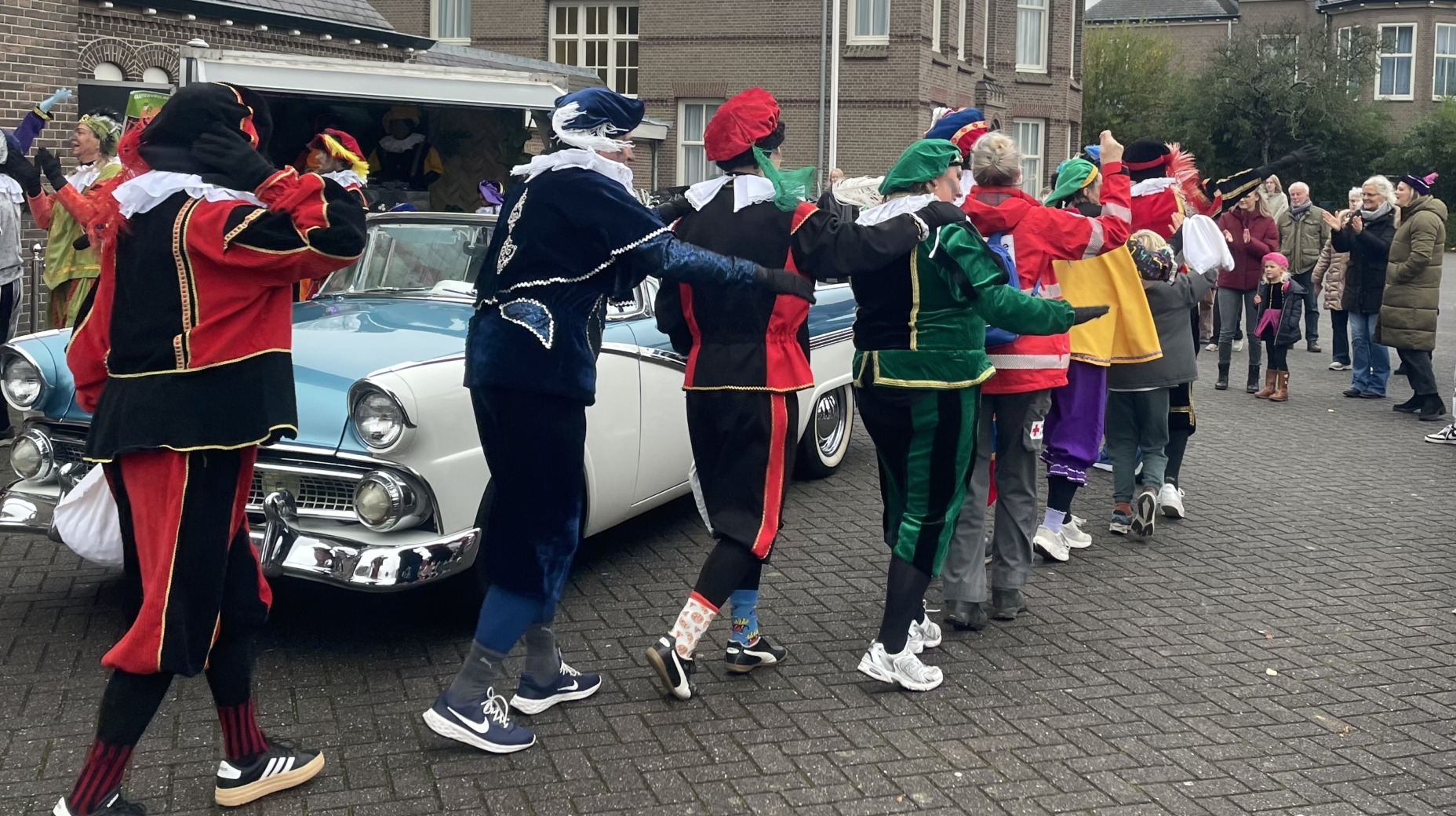 Een groep mensen in kleurrijke kleding danst in een polonaise voor een blauwe klassieke auto, omringd door toeschouwers bij een bakstenen gebouw.