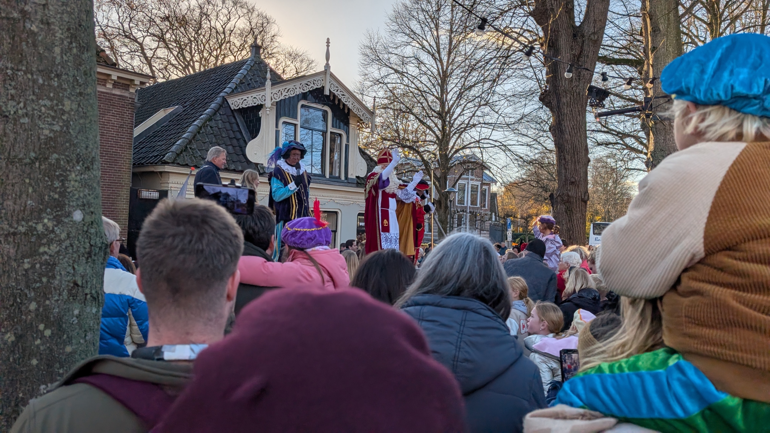 Sinterklaas en zijn helpers staan op een podium voor een menigte tijdens een optocht in een Nederlandse stad, omgeven door bomen en historische gebouwen.