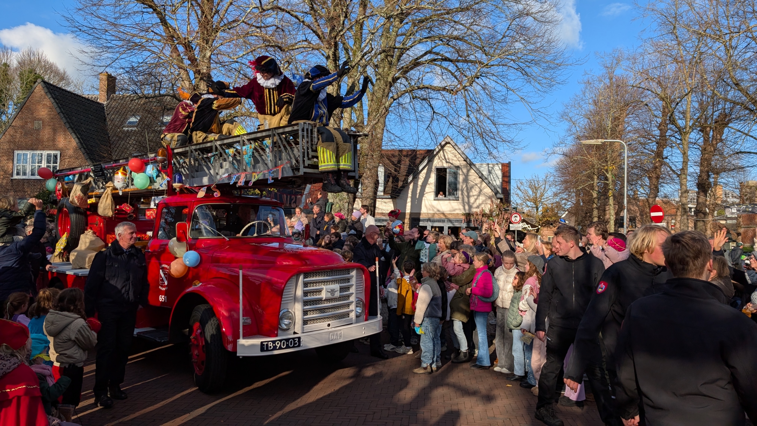 Een kleurrijke parade met een rode oude brandweerwagen waarop Pieten staan, die zwaaien naar een menigte van mensen, waaronder veel kinderen, op een zonnige dag in een bestraat dorp met bomen en huizen op de achtergrond.