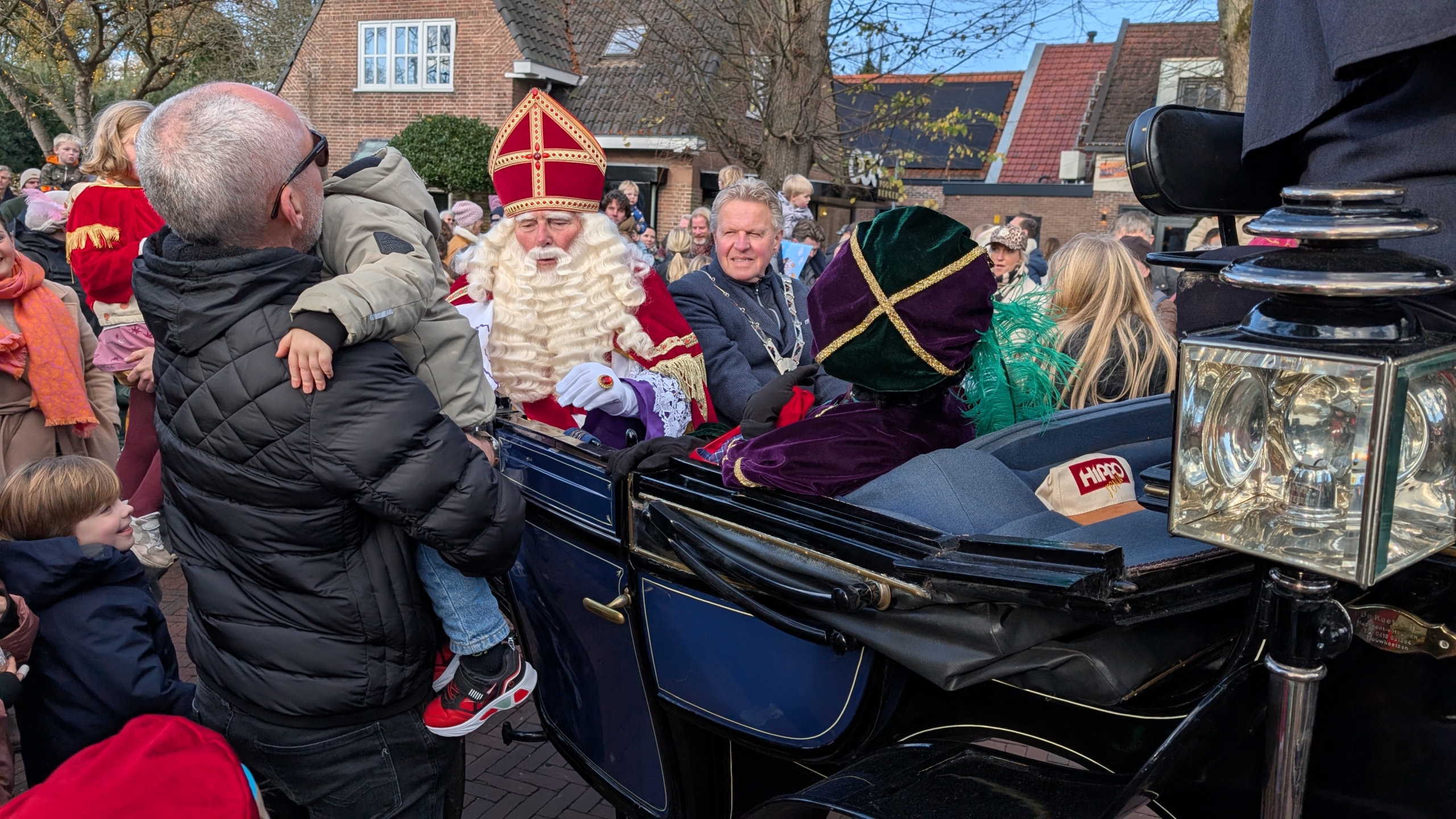 Sinterklaas en zijn Pieten zitten in een open koets tijdens een parade, omringd door lachende kinderen en volwassenen.
