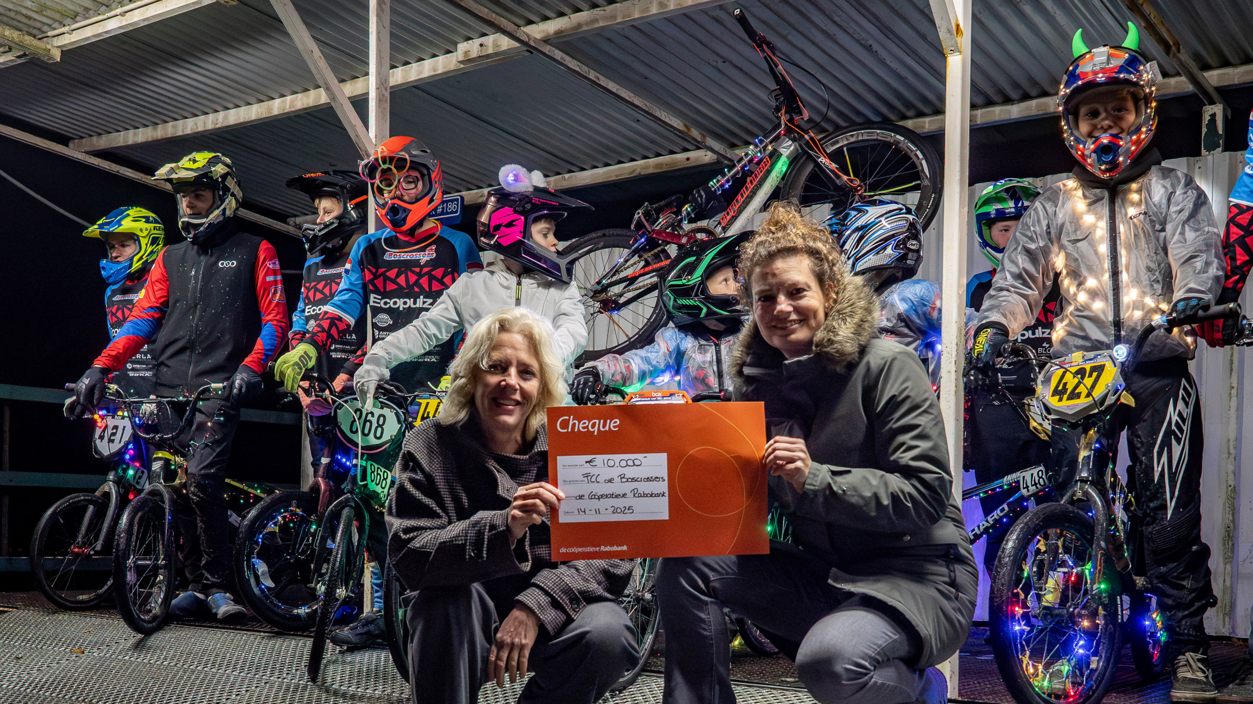Groep BMX-coureurs met kleurrijke en verlichte kleding en fietsen, geposeerd met twee volwassenen die een cheque van €10.000 vasthouden.