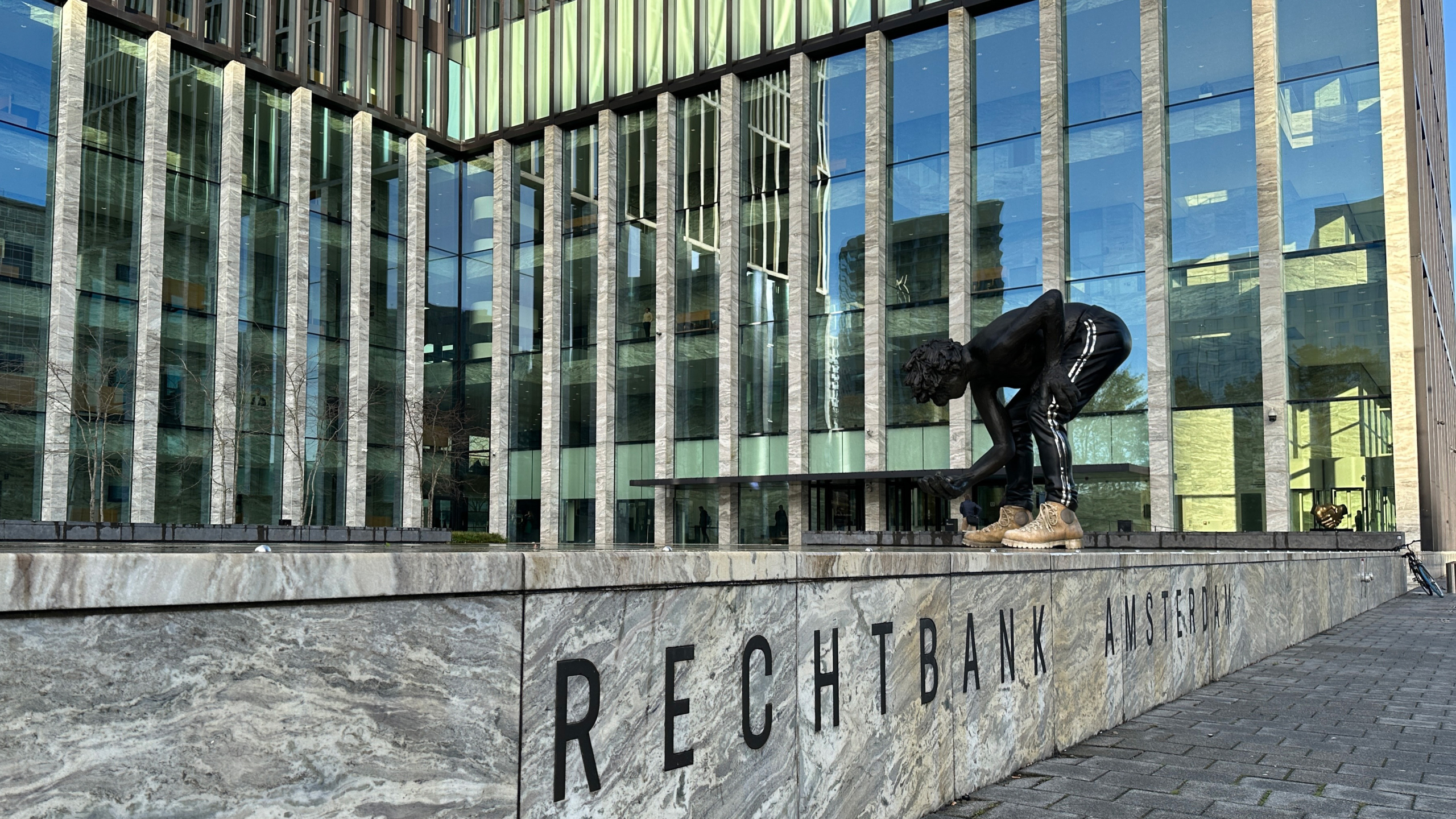 Buitenkant van een modern gebouw met glazen gevels en een groot standbeeld van een gebogen figuur in sportkleding en schoenen. Op de voorgrond staat de tekst "Rechtbank Amsterdam" op een stenen muur.