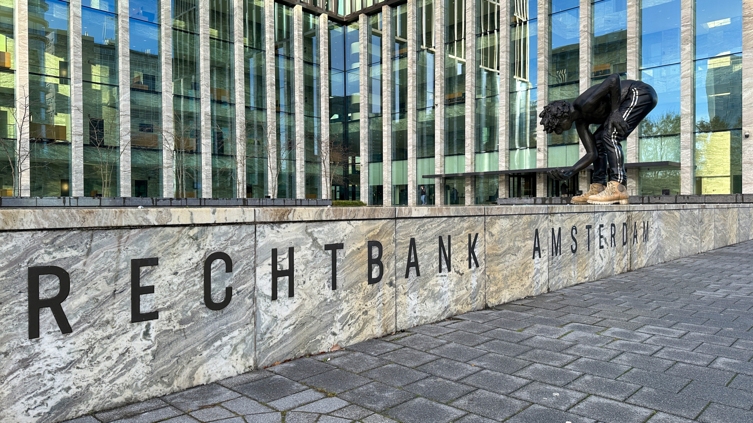 Buitenkant van de rechtbank in Amsterdam met een moderne glazen gevel en een standbeeld van een gebogen persoon, naast de zichtbare tekst 'RECHTBANK AMSTERDAM' op een marmeren muur.