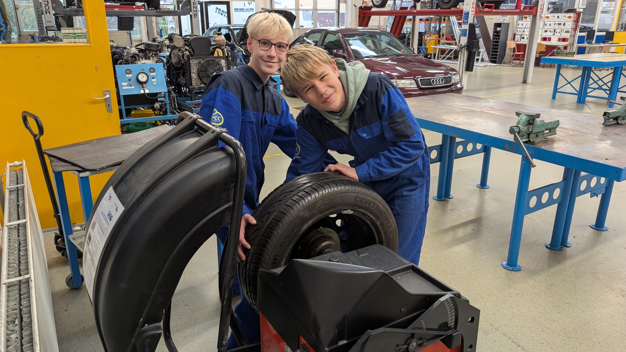 Twee jongens in blauwe overalls werken aan een autowiel in een werkplaats, met gereedschap en een voertuig op de achtergrond.