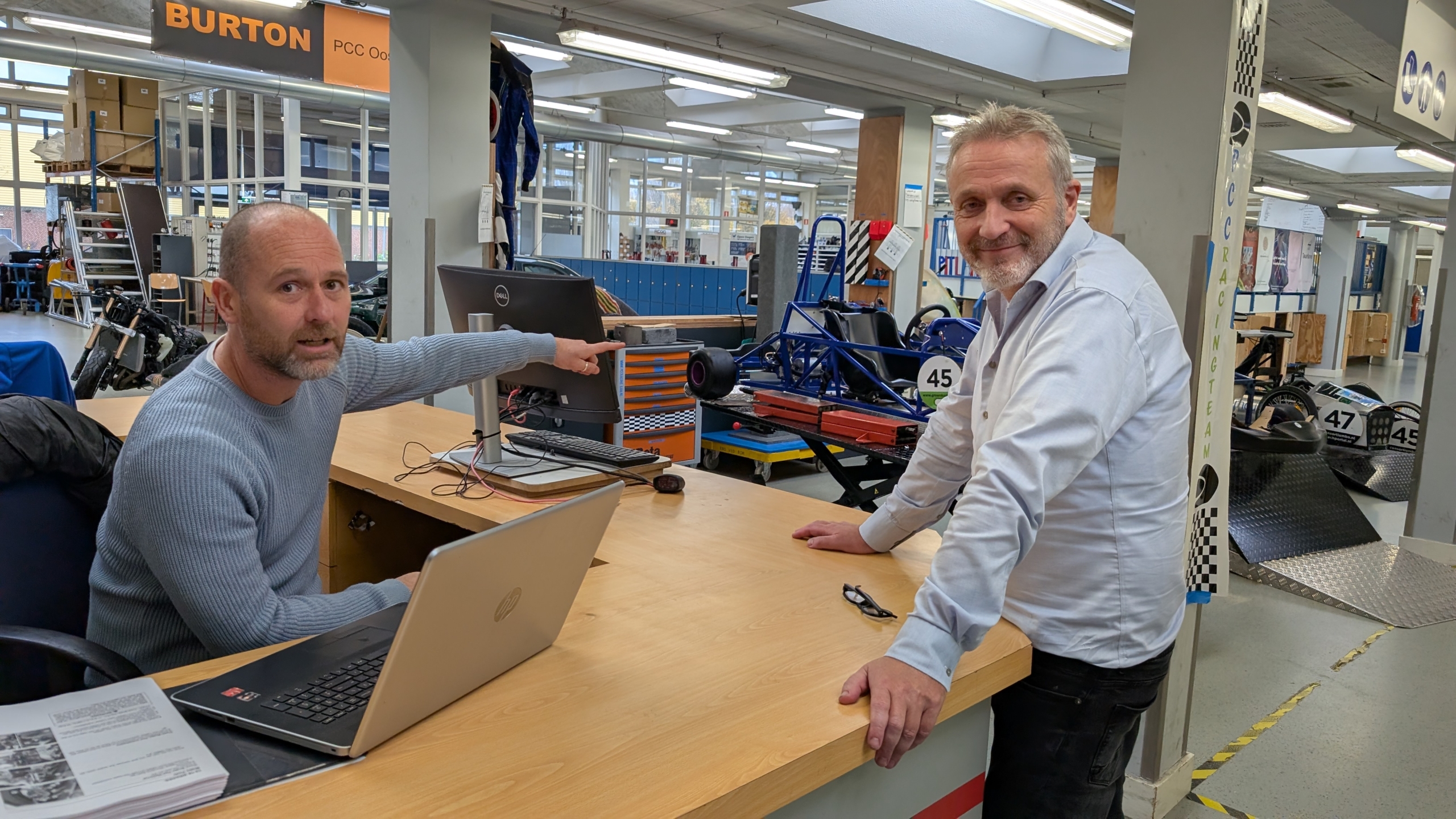 Twee mannen staan bij een balie in een werkplaats, met technische apparatuur en gedeeltelijk gemonteerde voertuigen op de achtergrond. Eén man wijst naar een computerscherm terwijl de andere glimlachend naar de camera kijkt.