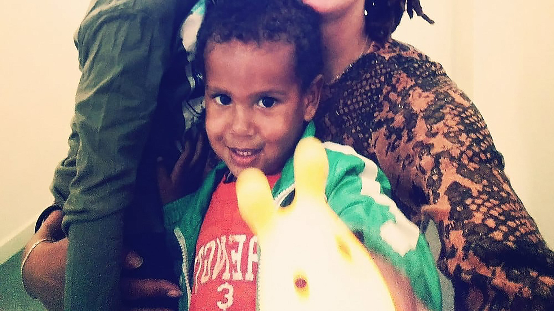 Een glimlachend kind in een gekleurde trui houdt een speeltje vast naast een volwassene in een bruinig patroon shirt.