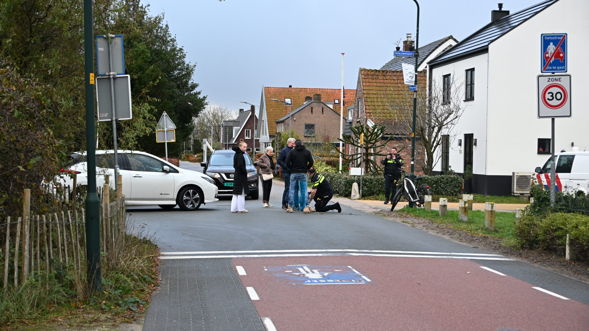 Groep mensen bij een straat met politie, auto's en verkeersborden in Nederland, terwijl een agent iemand helpt op de grond.