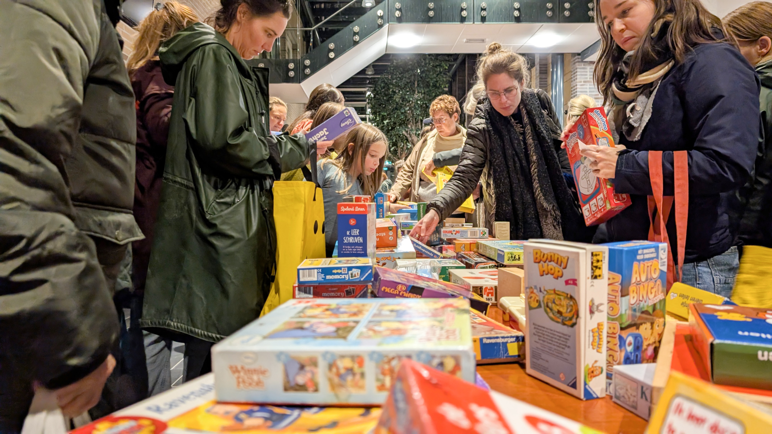 Mensen verzamelen zich rond een tafel met diverse bordspellen, terwijl ze door de spellen bladeren en enkele spellen vasthouden in een binnenruimte.