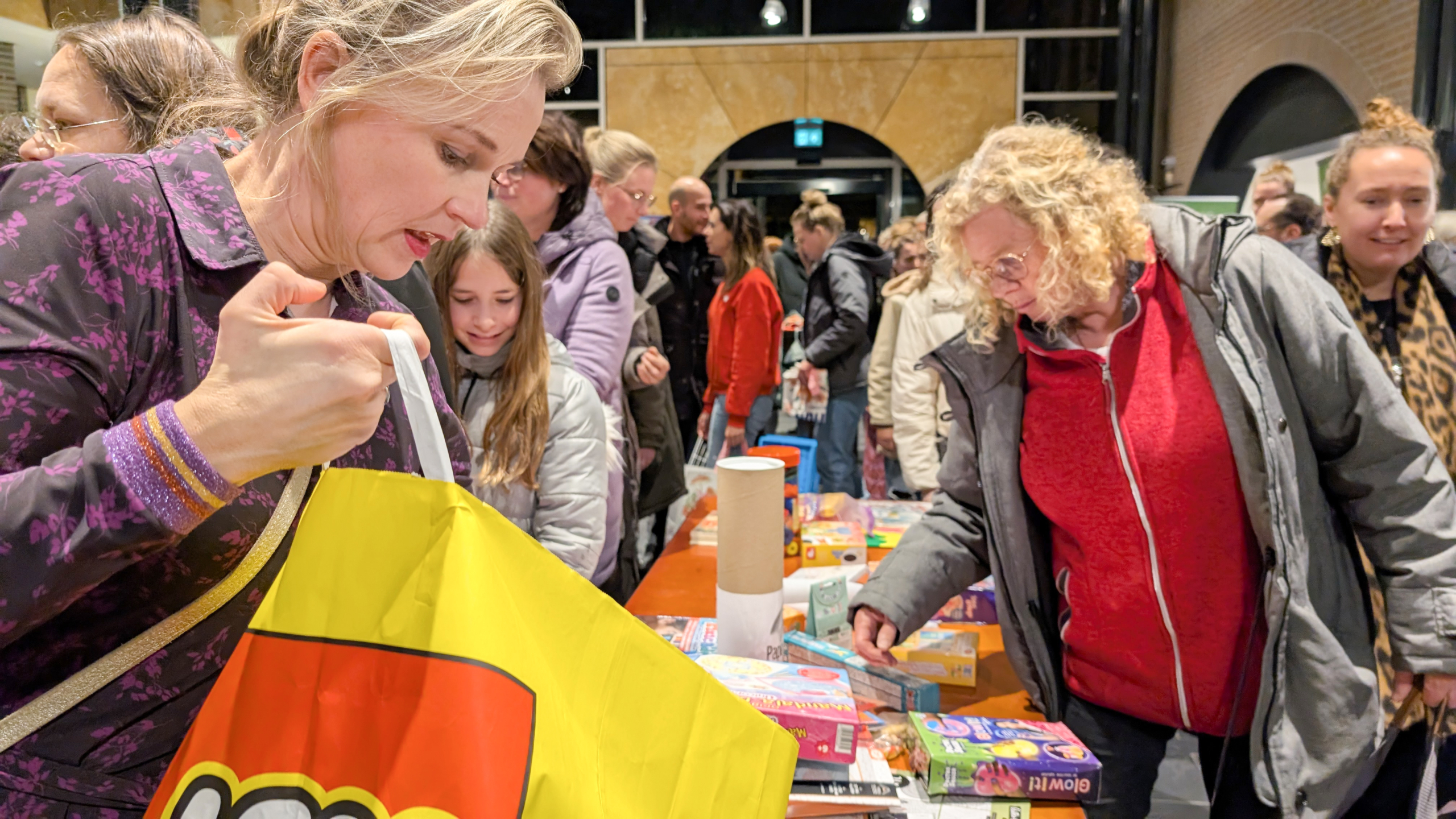 Mensen zoeken naar speelgoed op een tafel tijdens een druk bezochte bijeenkomst, waarbij een vrouw de inhoud bekijkt van een grote geel-rode tas met LEGO-logo.